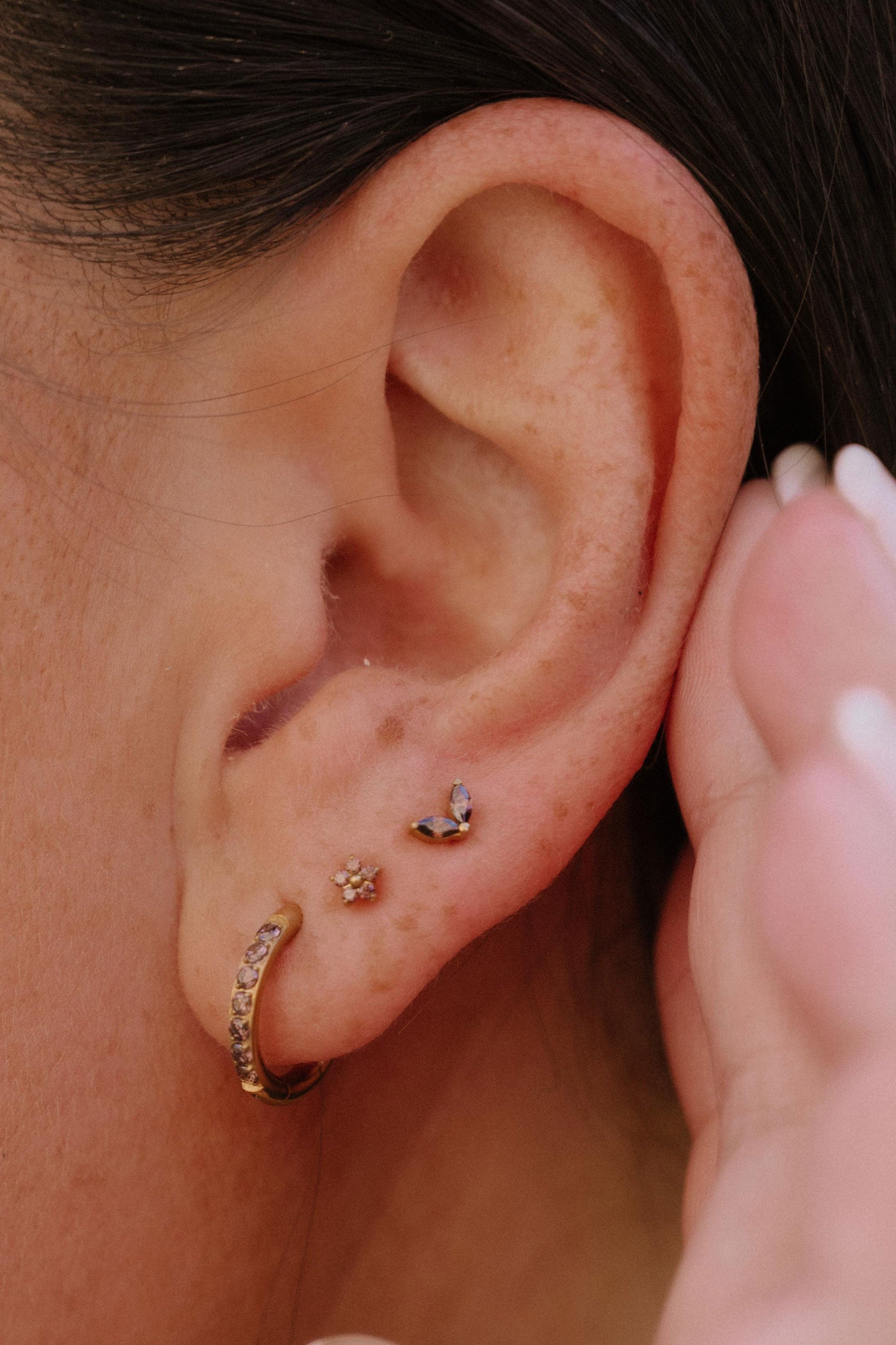 Close-up of a person's ear showcasing multiple small gold piercings, including a hoop, a flower stud, and a double leaf-shaped stud.