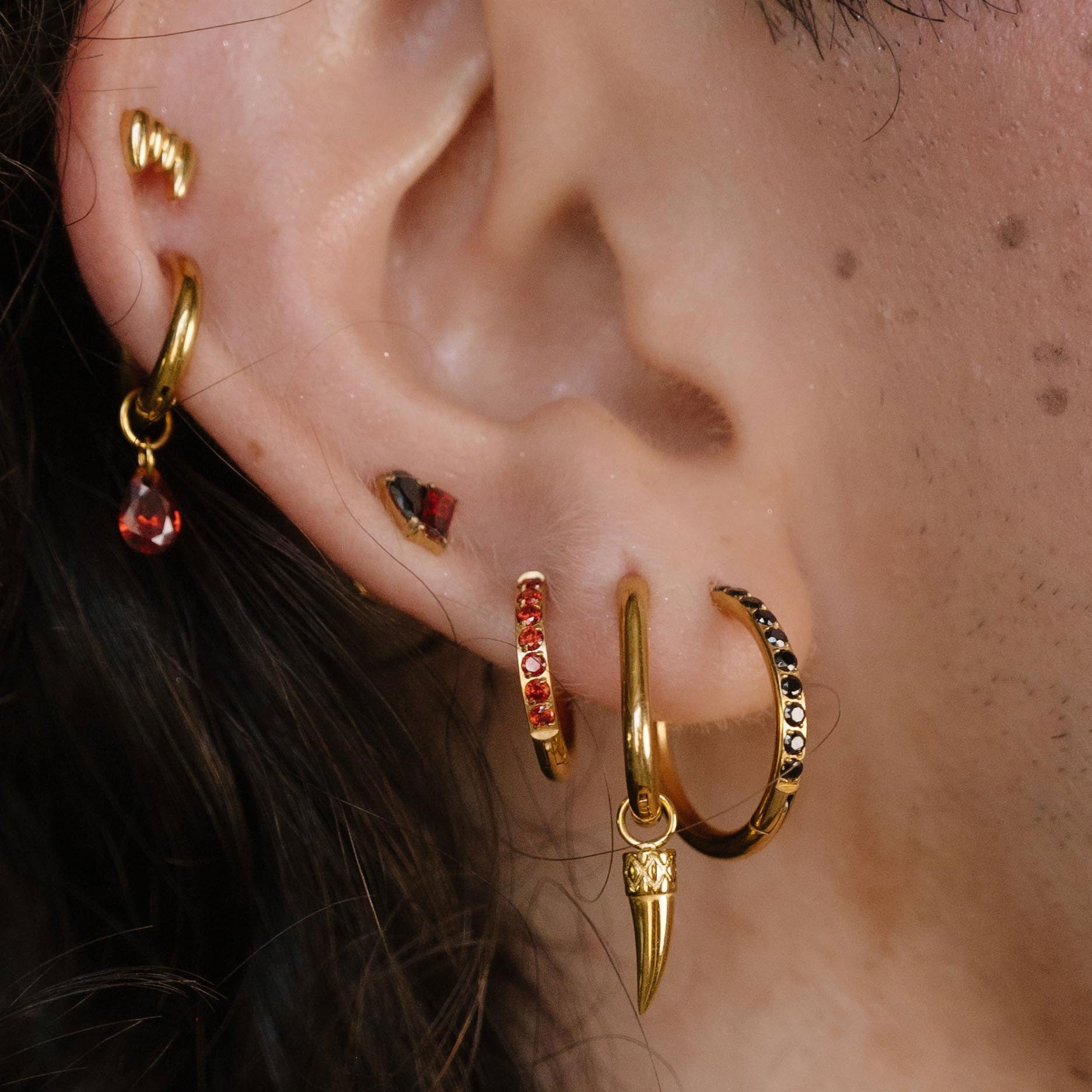 Close-up of a person's ear wearing multiple gold hoop and cartilage earrings, with small red gemstone accents and a gold leaf-shaped earring, against dark curly hair and a gold chain necklace.