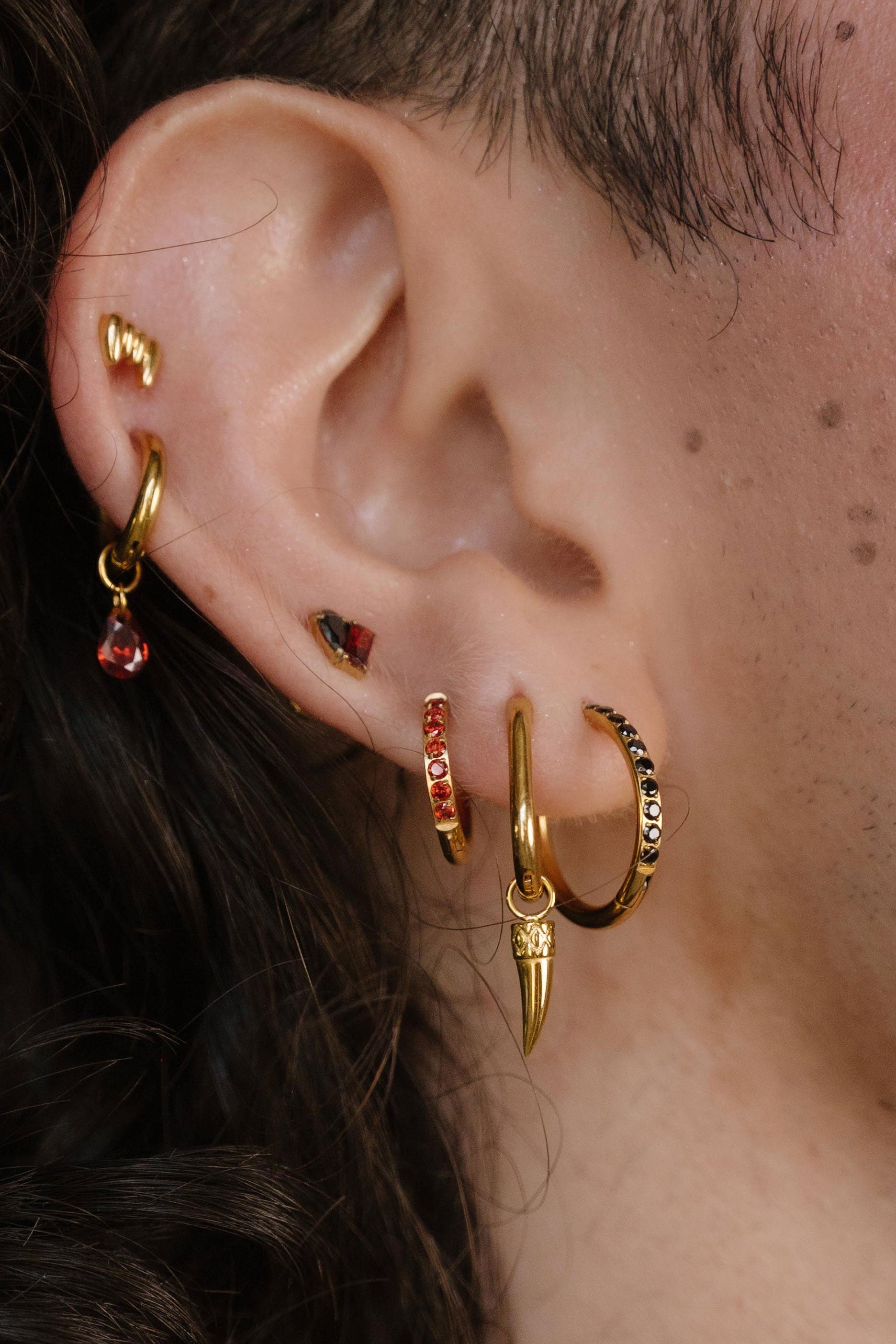 Close-up of a person's ear wearing multiple gold hoop and cartilage earrings, with small red gemstone accents and a gold leaf-shaped earring, against dark curly hair and a gold chain necklace.