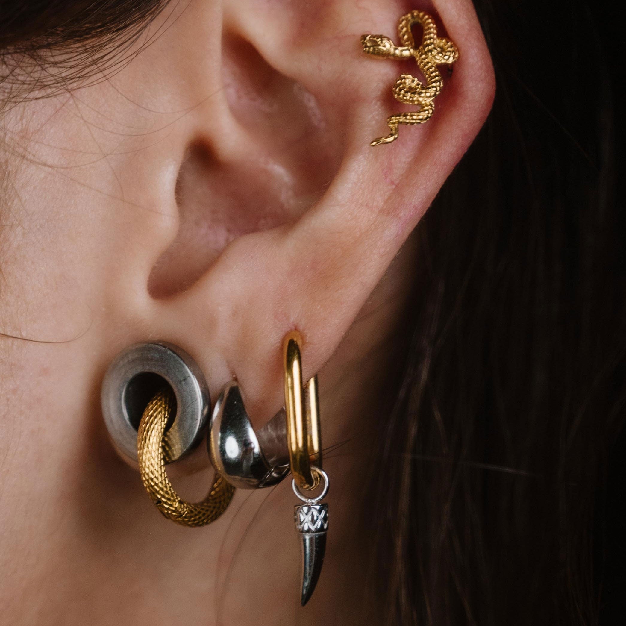 Close-up of a person's ear showcasing multiple stacked and cartilage piercings, including gold hoop, silver and gold cartilage studs, and a small silver drop with a clear stone.