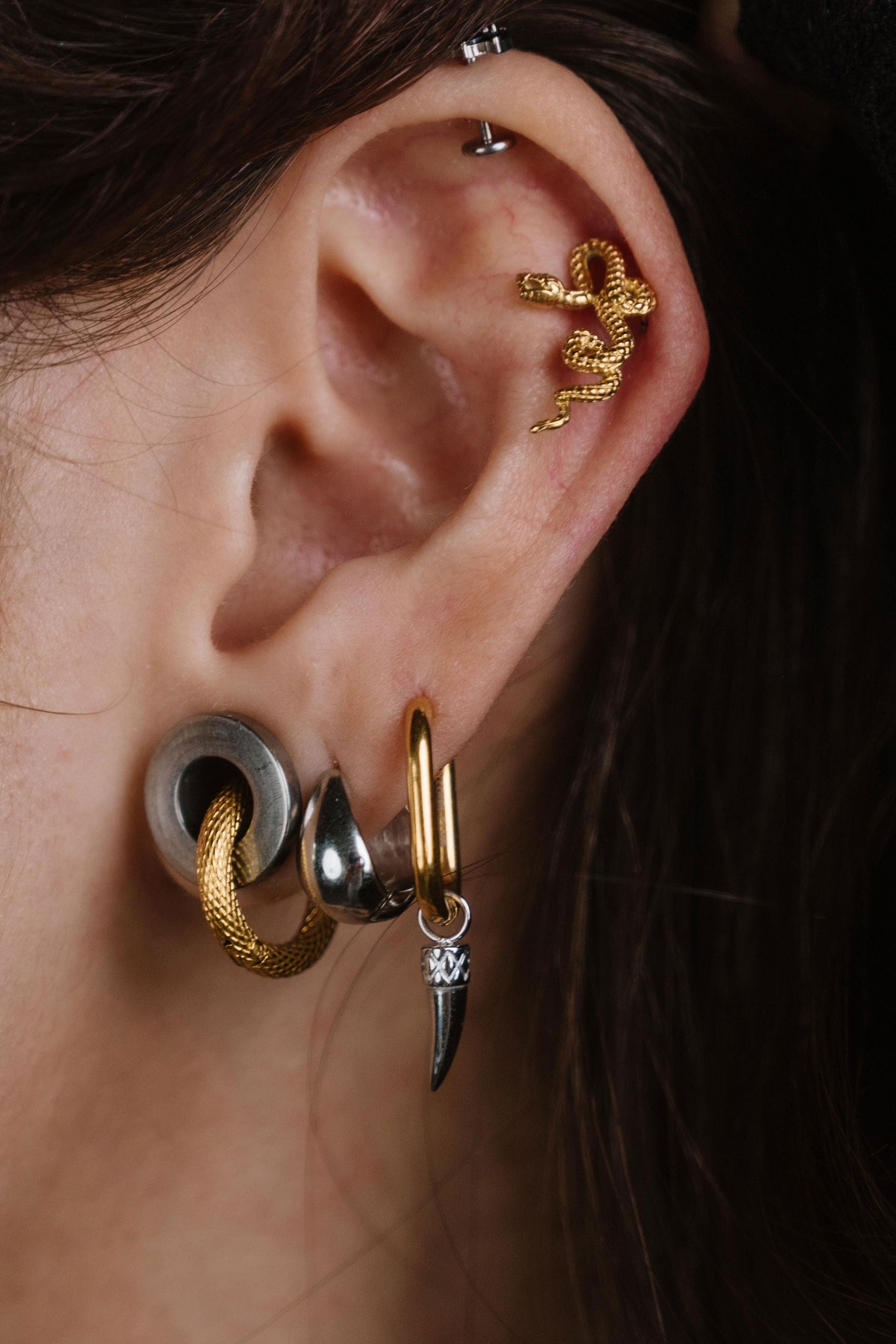 Close-up of a person's ear showcasing multiple stacked and cartilage piercings, including gold hoop, silver and gold cartilage studs, and a small silver drop with a clear stone.