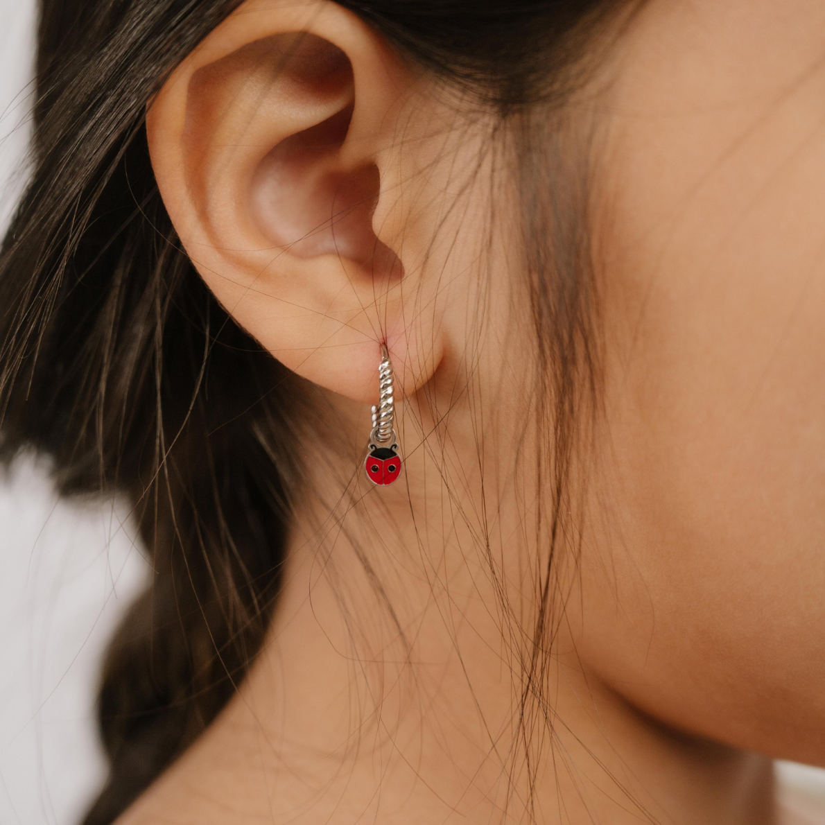 A close up of a ladybug shaped hoop charm with red and black enamel and  titanium metal hanging from a hoop earring.