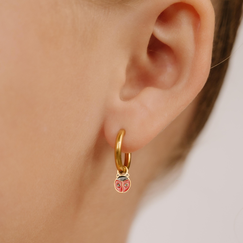 A close up of a ladybug shaped hoop charm with red and black enamel and gold-plated titanium metal hanging from a hoop earring.