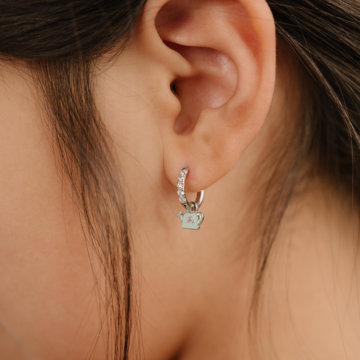 A close up of a right-facing watering can shaped charm with mint green enamel and titanium metal hanging from a hoop earring.