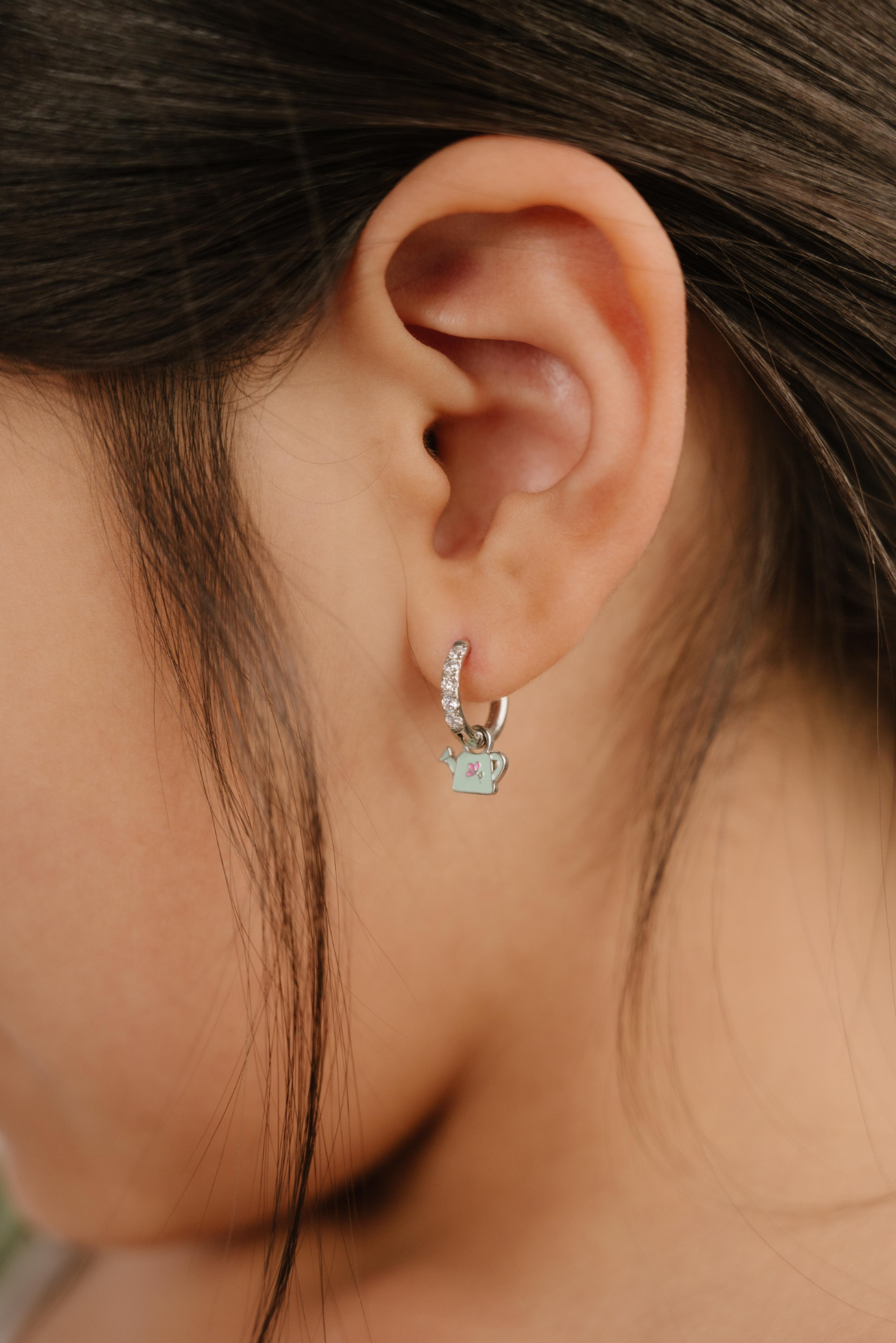 A close up of a right-facing watering can shaped charm with mint green enamel and titanium metal hanging from a hoop earring.