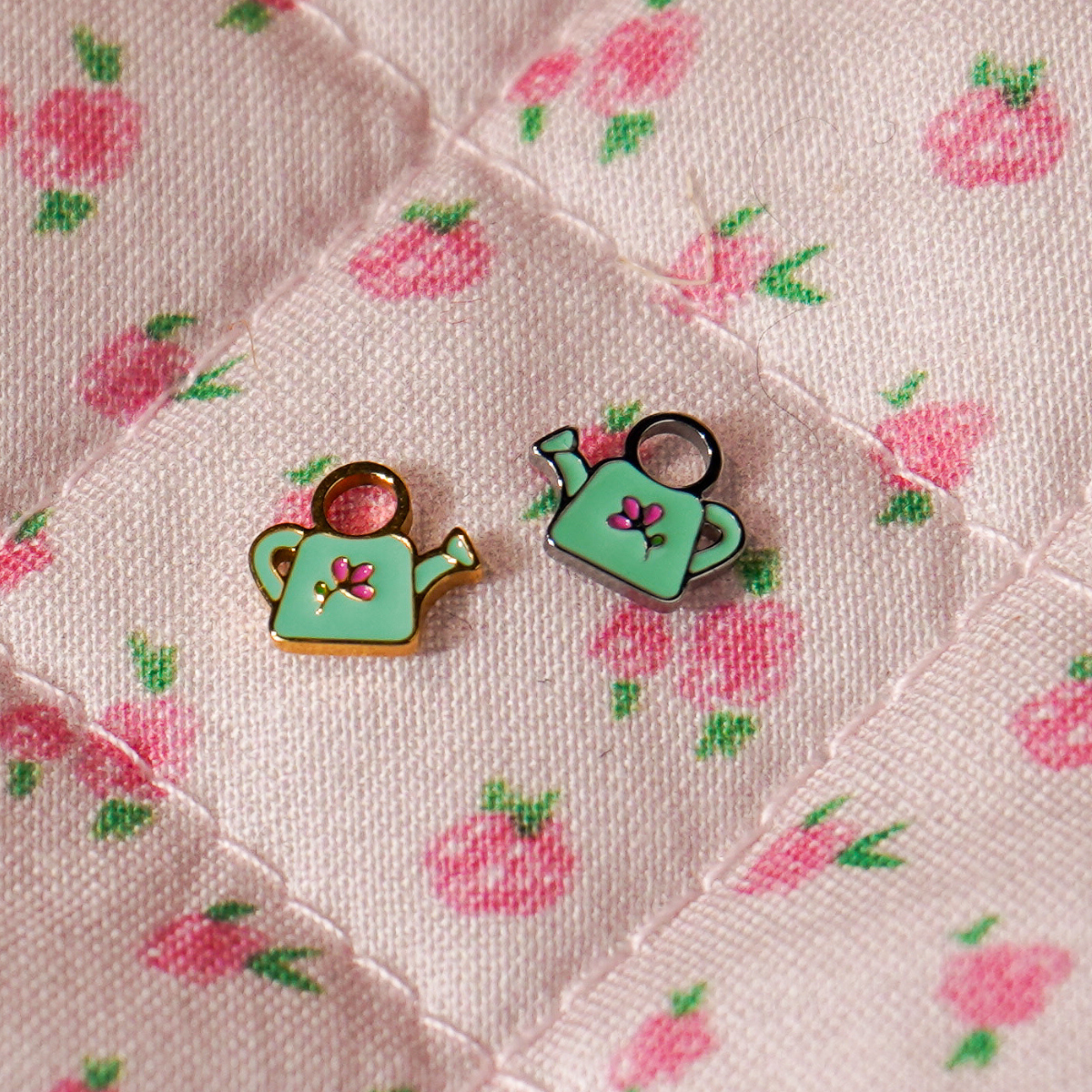 A close up of 2 watering can shaped hoop charms with mint green enamel and titanium metal on a pink floral background