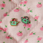 A close up of 2 watering can shaped hoop charms with mint green enamel and titanium metal on a pink floral background