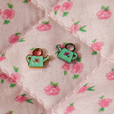 A close up of 2 watering can shaped hoop charms with mint green enamel and titanium metal on a pink floral background