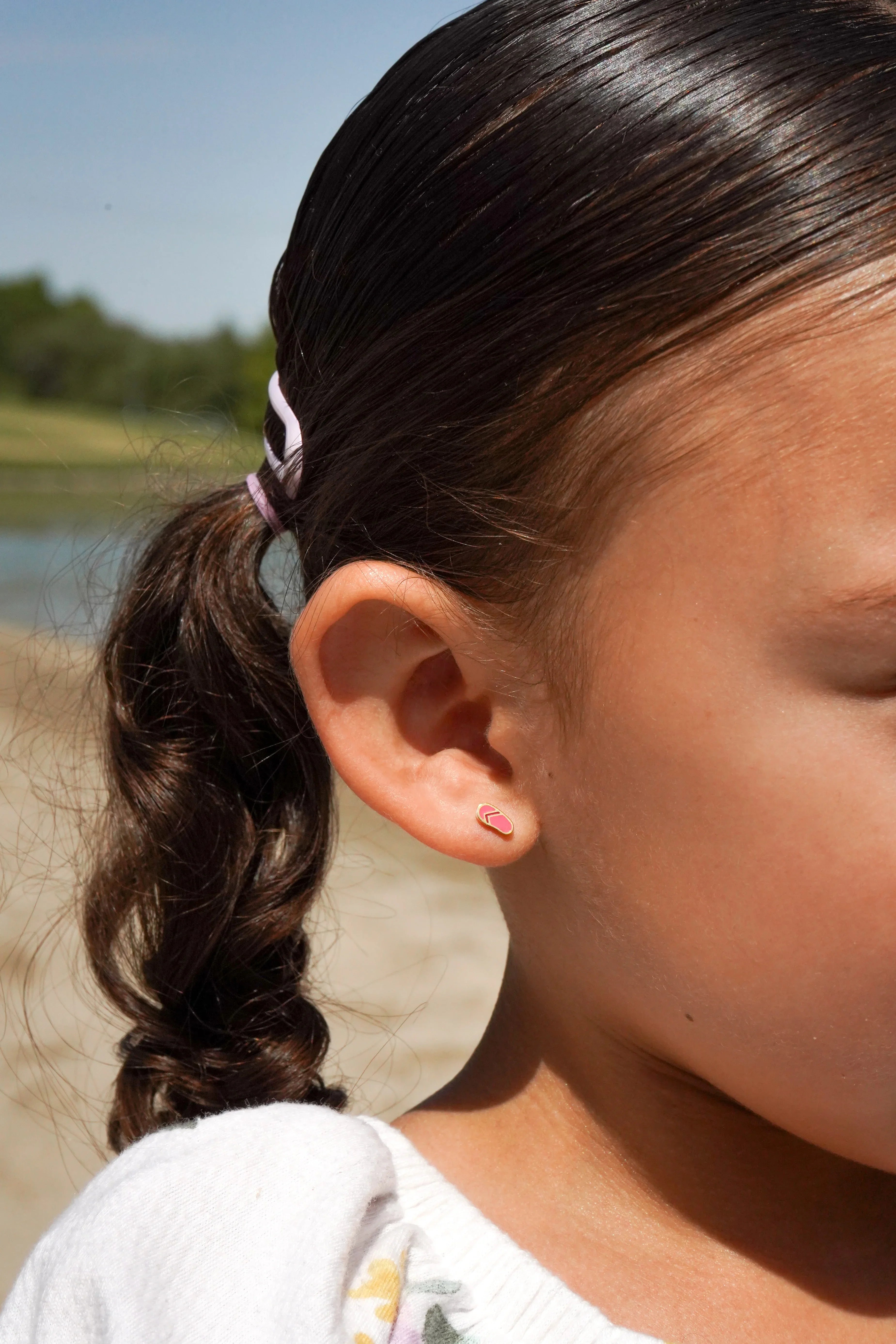 Close-up of a young girl with a curly ponytail and pink hair tie, and a pink sandal-shaped earring, outdoors.