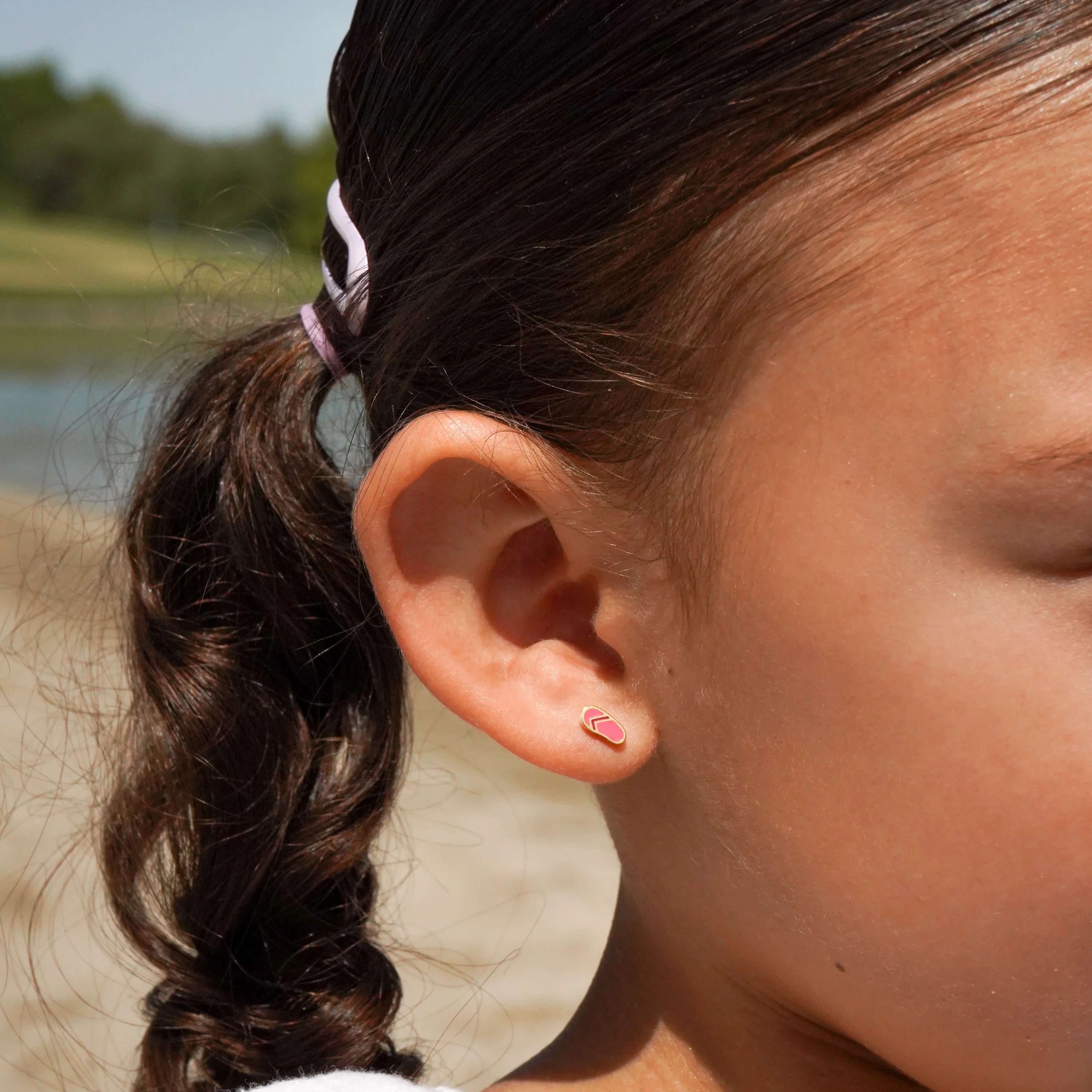 Close-up of a young girl with a curly ponytail and pink hair tie, and a pink sandal-shaped earring, outdoors.