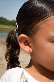 Close-up of a young girl with a curly ponytail and pink hair tie, and a pink sandal-shaped earring, outdoors.