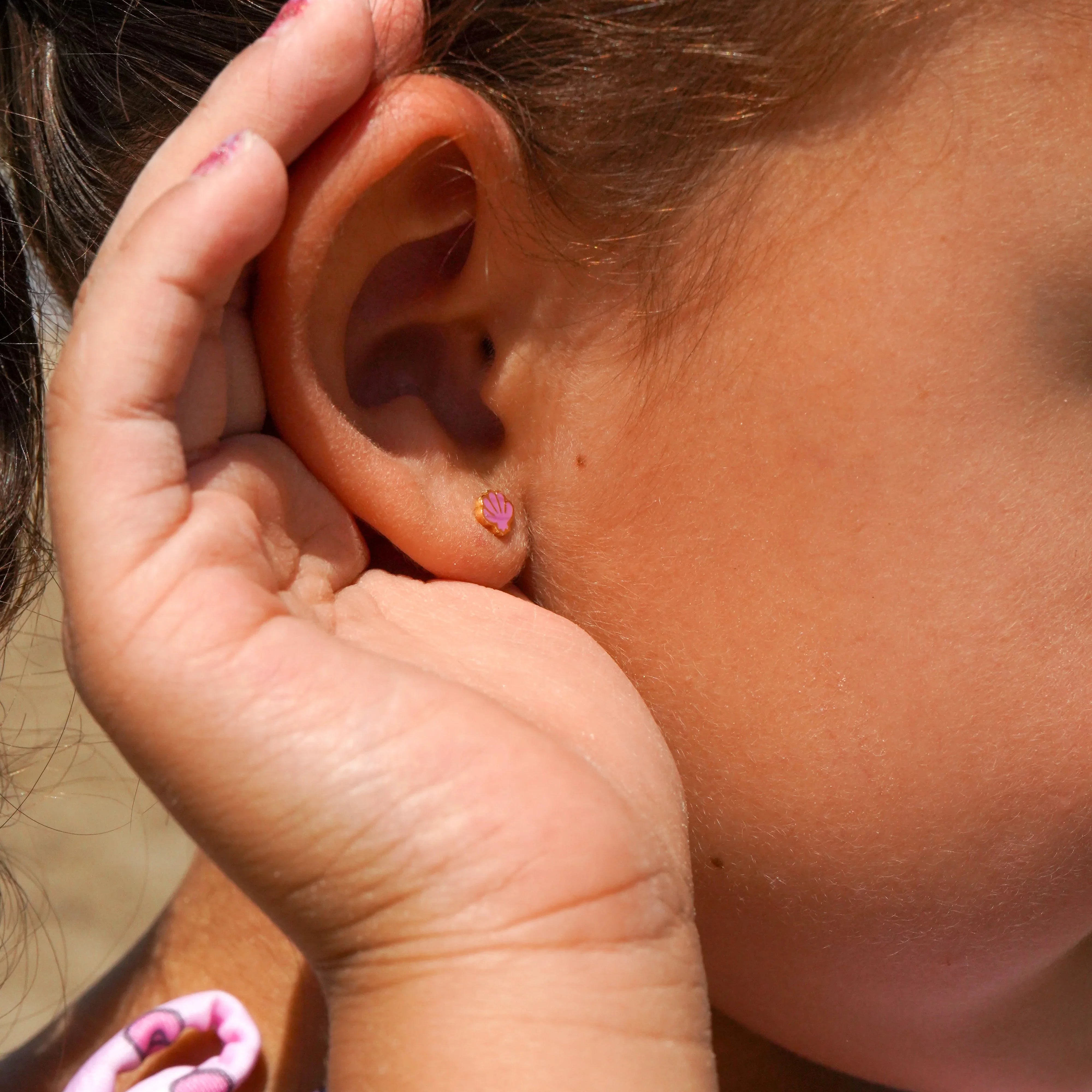 Close-up of a child's hand holding their ear, showcasing a purple shell earring.