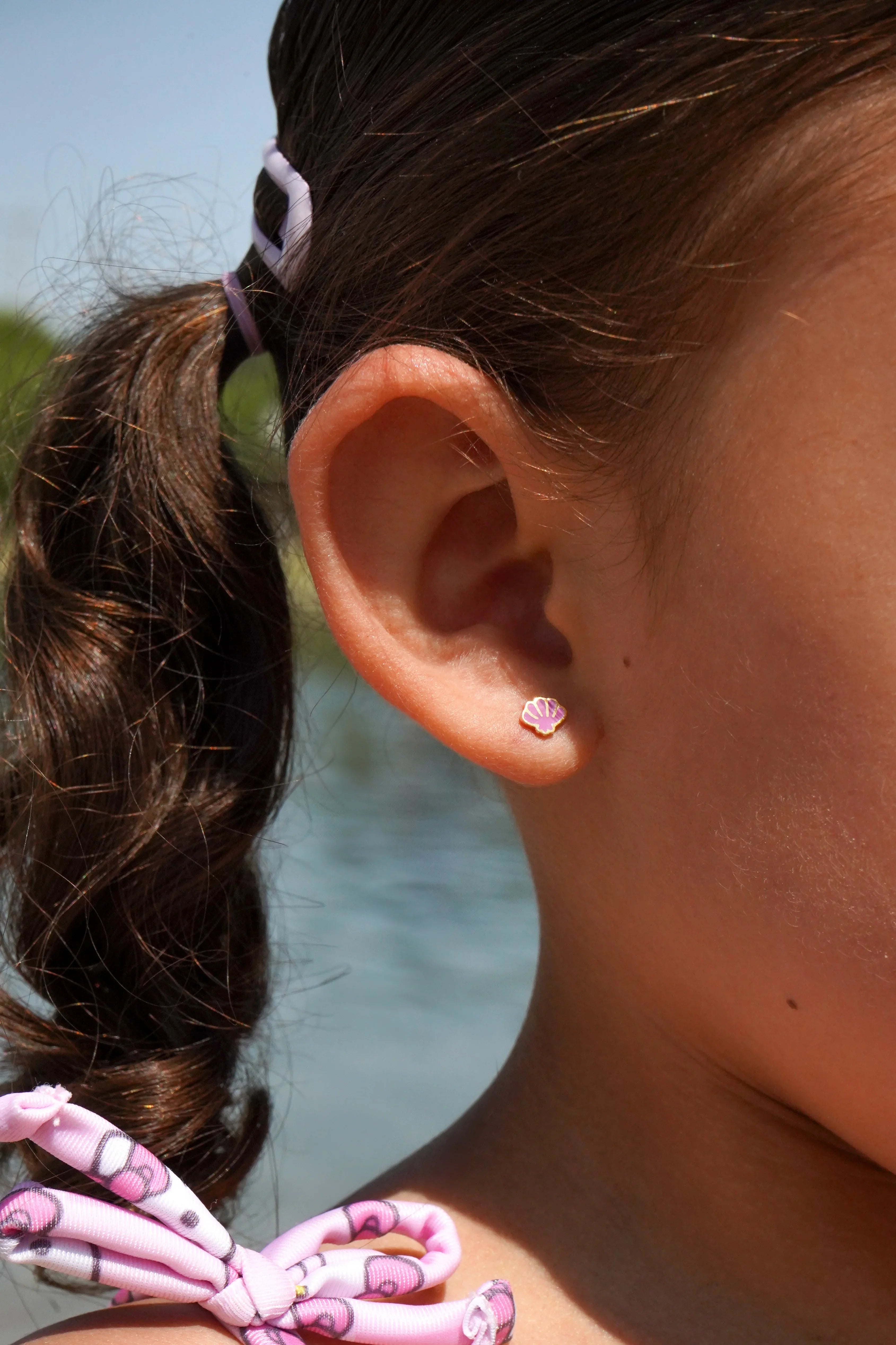 Close-up of a child's ear with a purple shell-shaped earring, blurred natural background