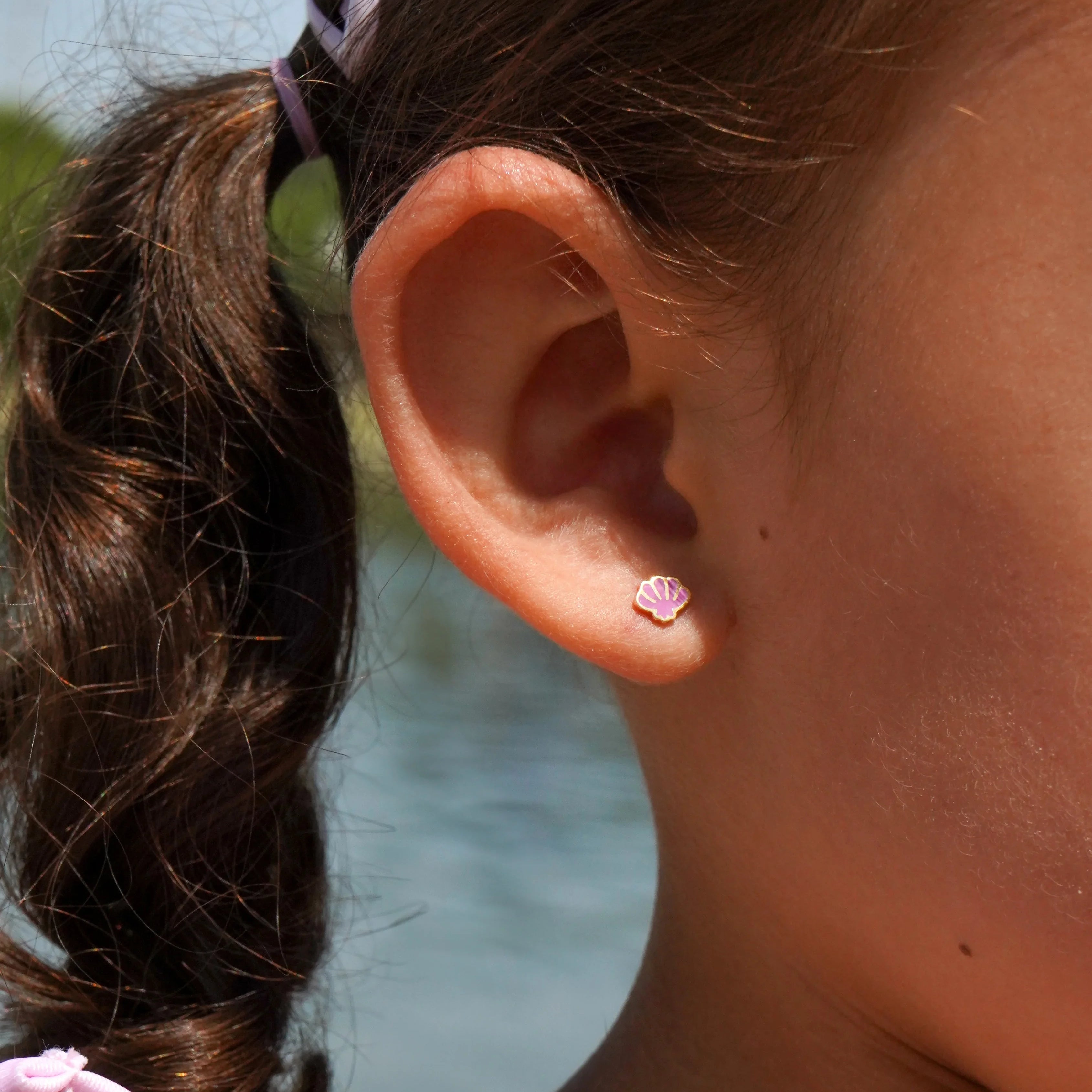 Close-up of a child's ear with a purple shell-shaped earring, blurred natural background