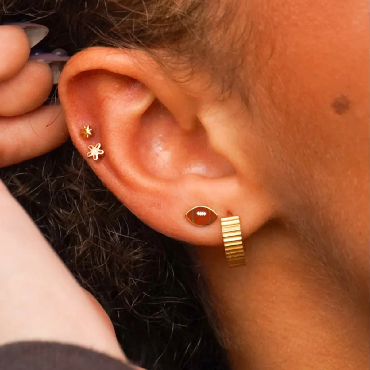 Close-up of an ear with gold hoop earrings and a small stud, with a hand adjusting something near the ear.