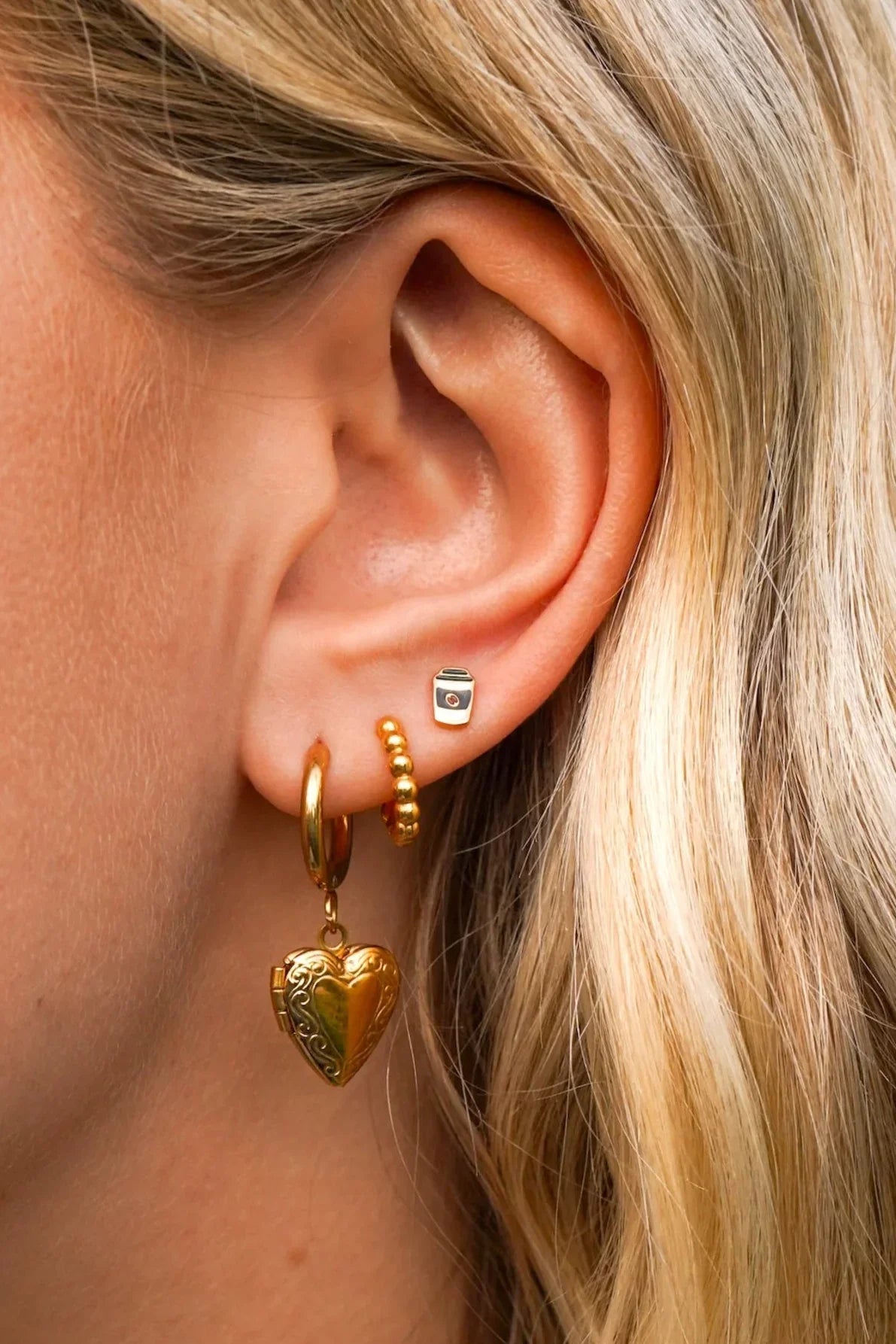 Close-up of an ear with gold hoop earrings, a heart-shaped earring, and an enamel coffee cup-shaped stud.