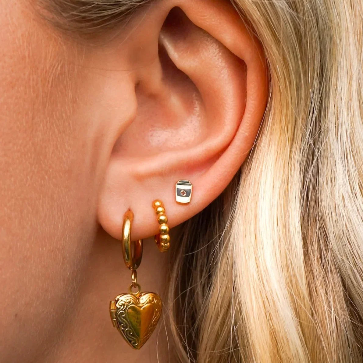 Close-up of an ear with gold hoop earrings, a heart-shaped earring, and an enamel coffee cup-shaped stud.