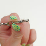 A close-up of a hand with green and yellow patterned nail art holding a silver chain and a side view of a silver compass pendant engraved with 'FOLLOW YOUR TRUTH'.