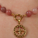 A close-up of a beaded necklace worn on a person's neck, featuring round pinkish-red beads and a gold compass pendant.