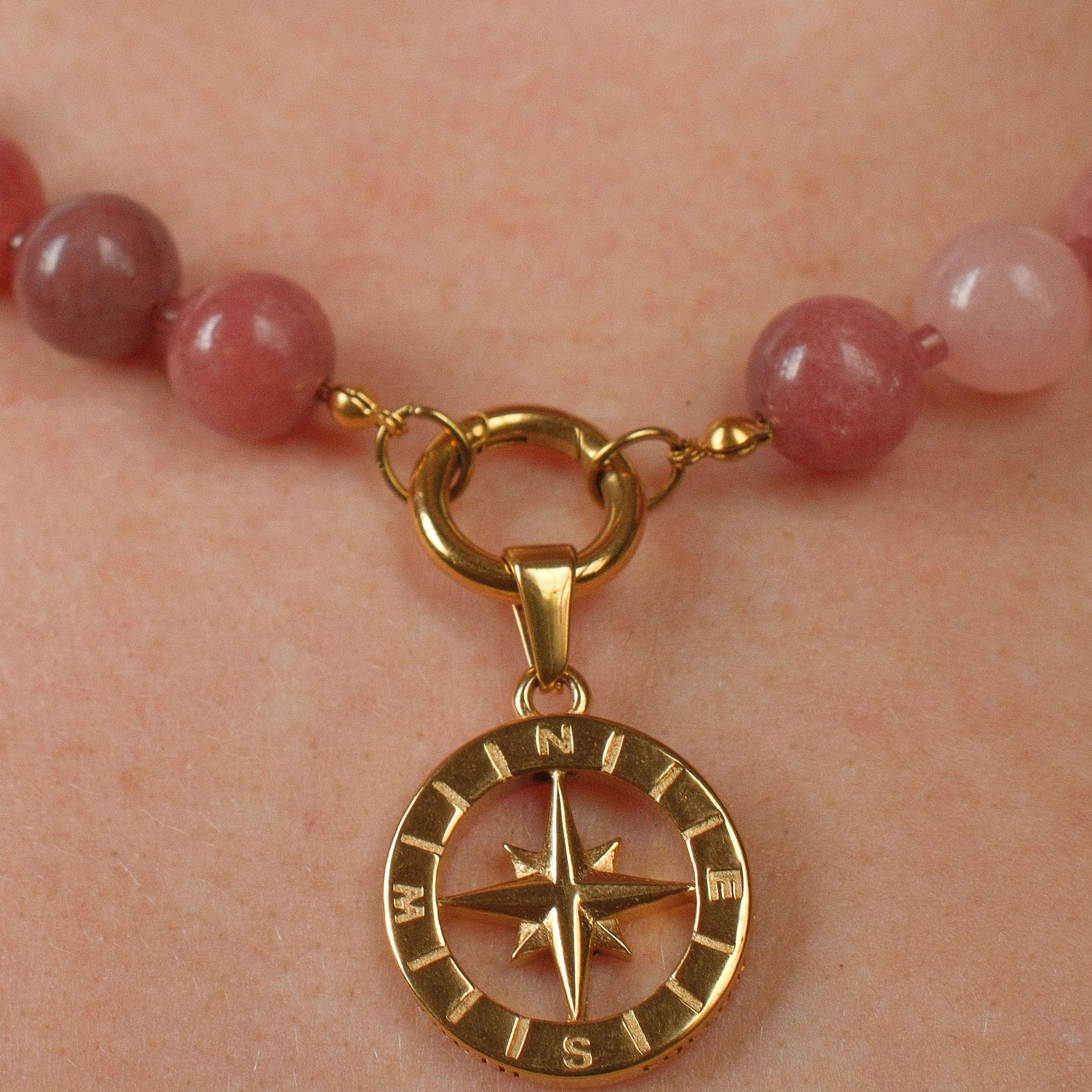 A close-up of a beaded necklace worn on a person's neck, featuring round pinkish-red beads and a gold compass pendant.