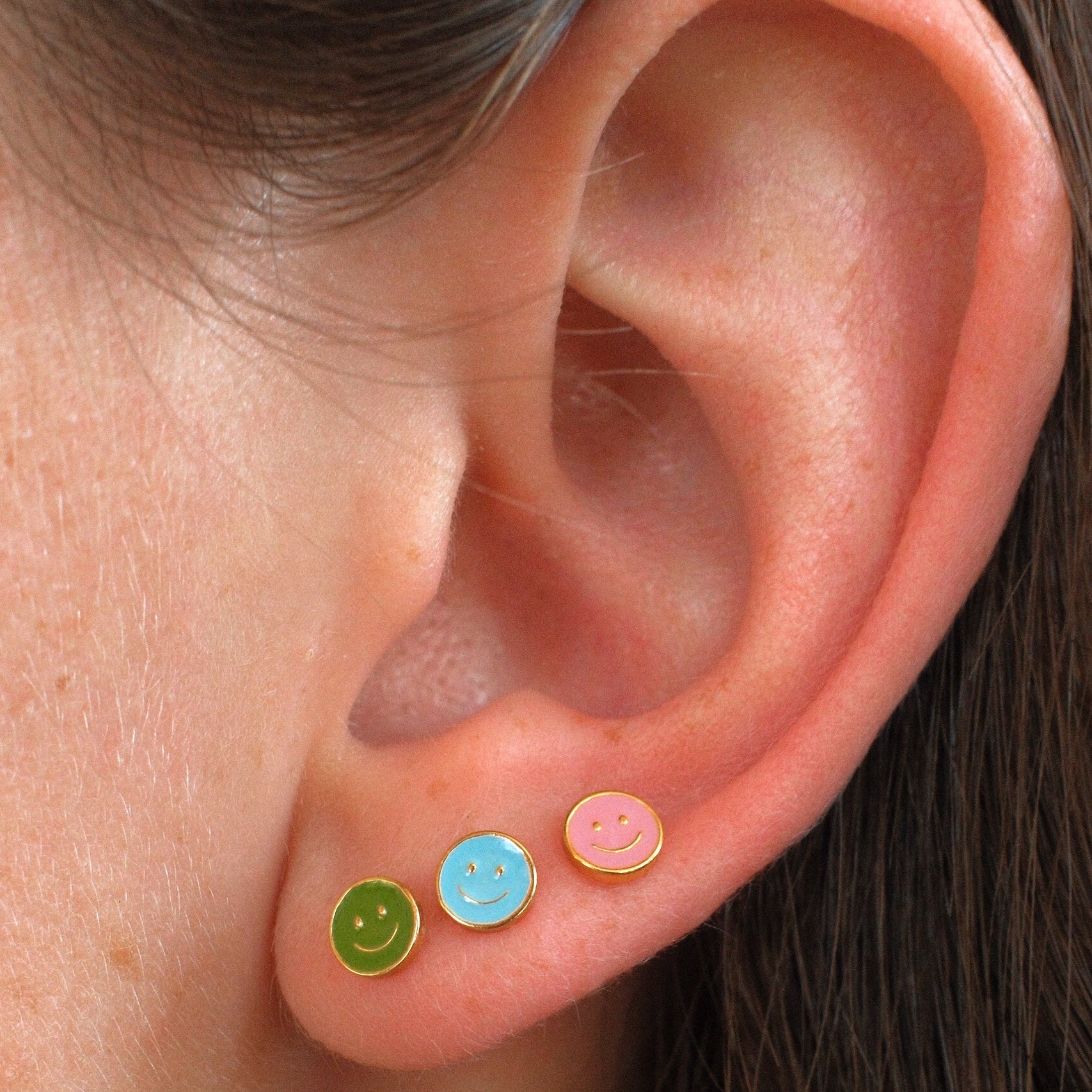 Close-up of a person's ear wearing three small smiley face stud earrings in green, light blue, and pink.
