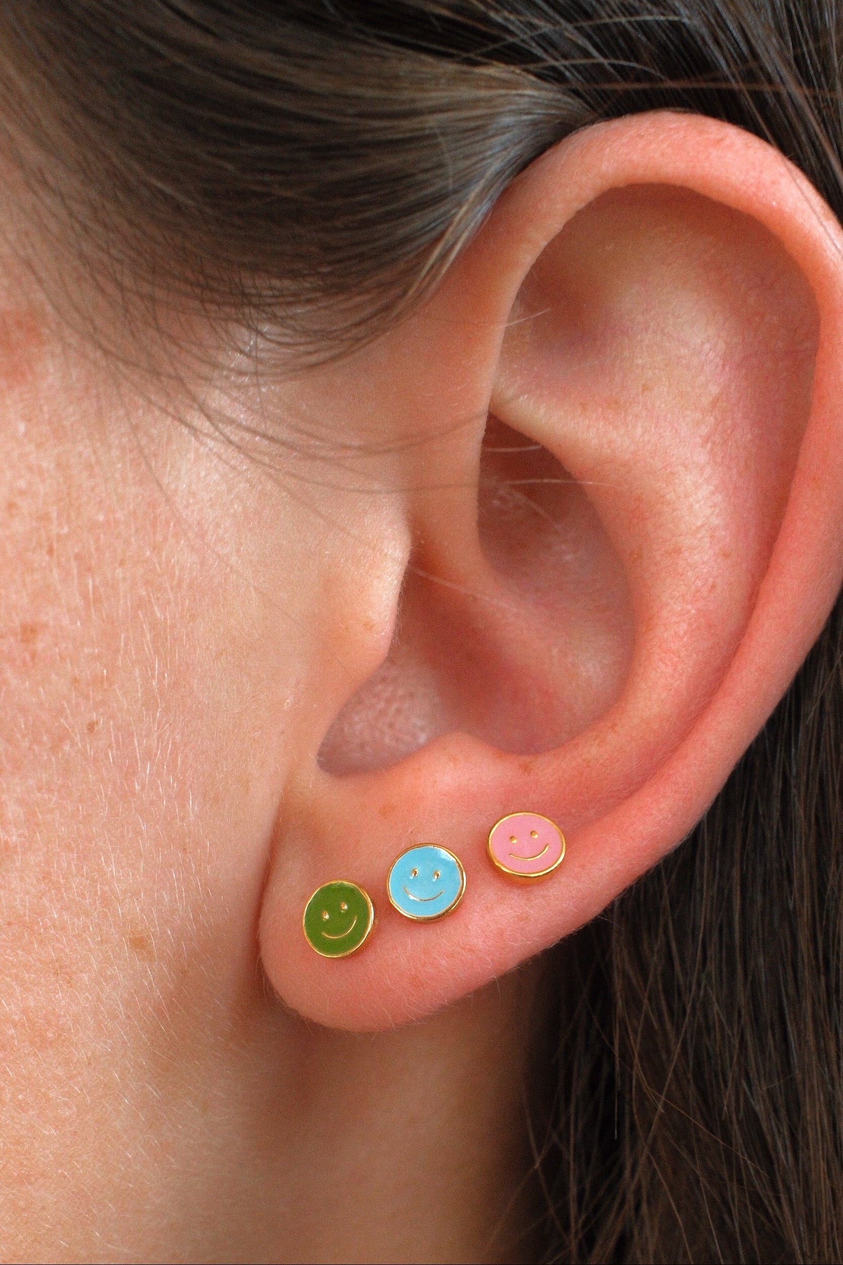 Close-up of a person's ear wearing three small smiley face stud earrings in green, light blue, and pink.