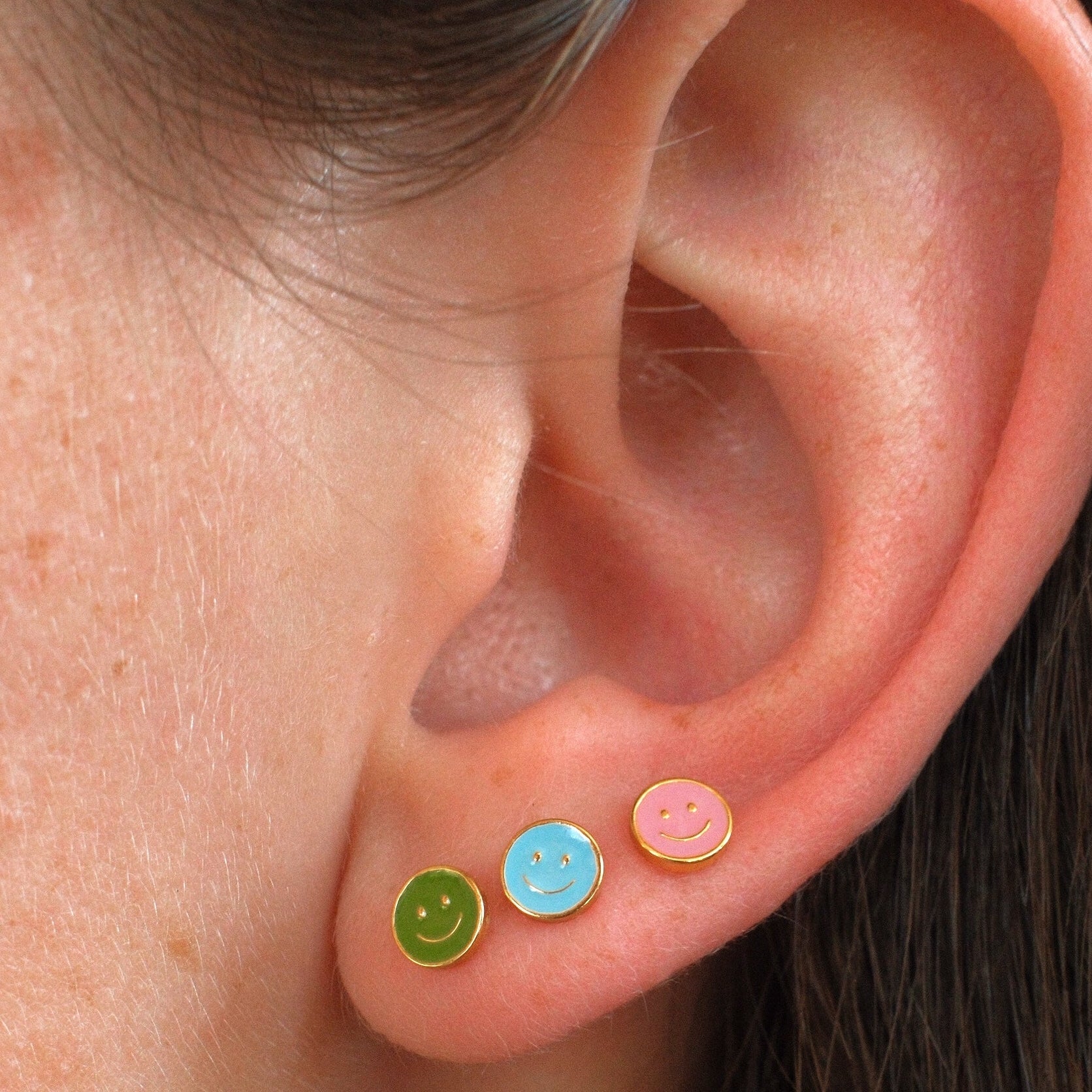 Close-up of a person's ear wearing three small smiley face stud earrings in green, light blue, and pink.