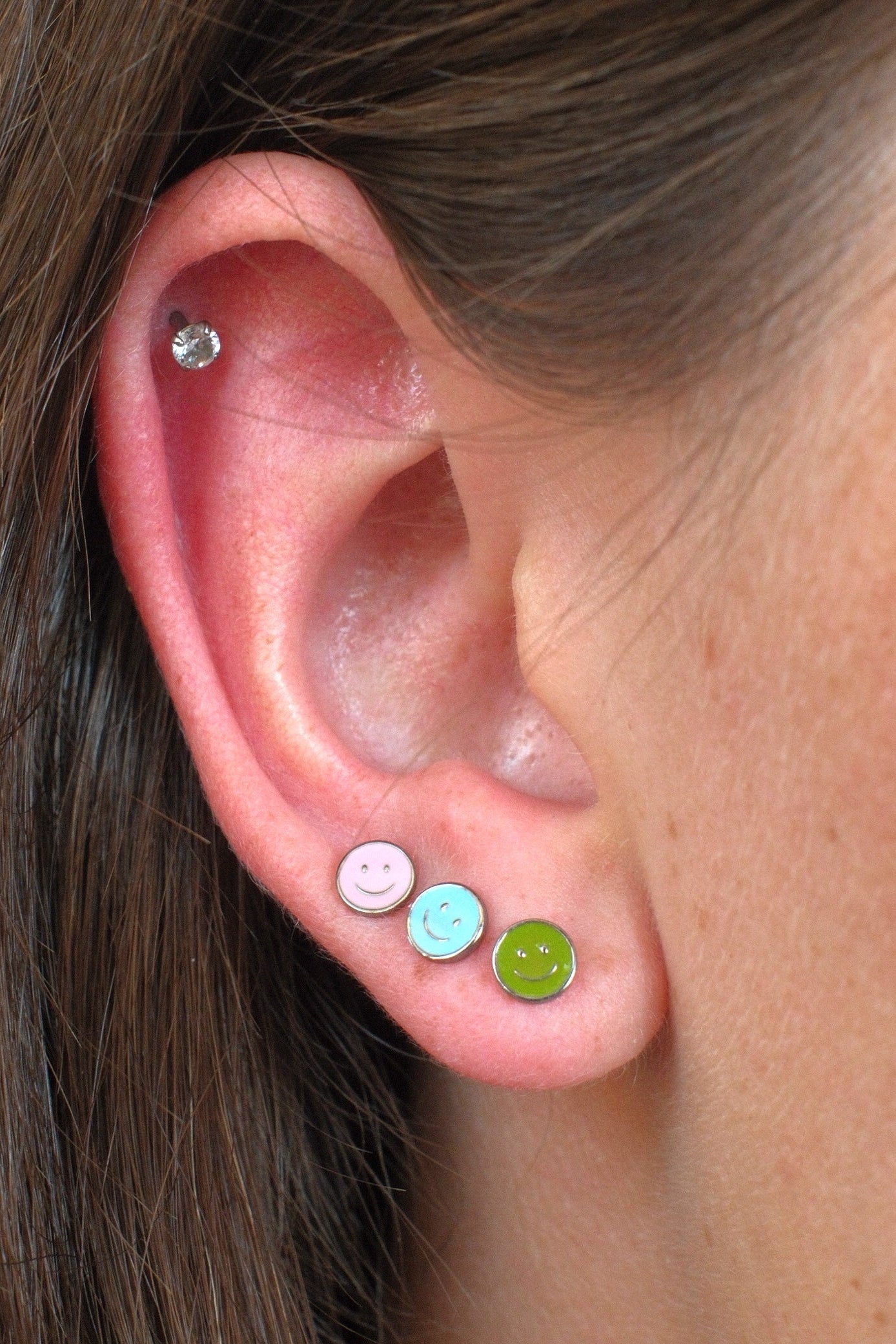 Close-up of a person's ear showcasing three colorful stud earrings: a pink smiley face stud, a light blue smiley face stud, and a lime green smiley face stud, with a small silver hoop earring in the upper ear cartilage.