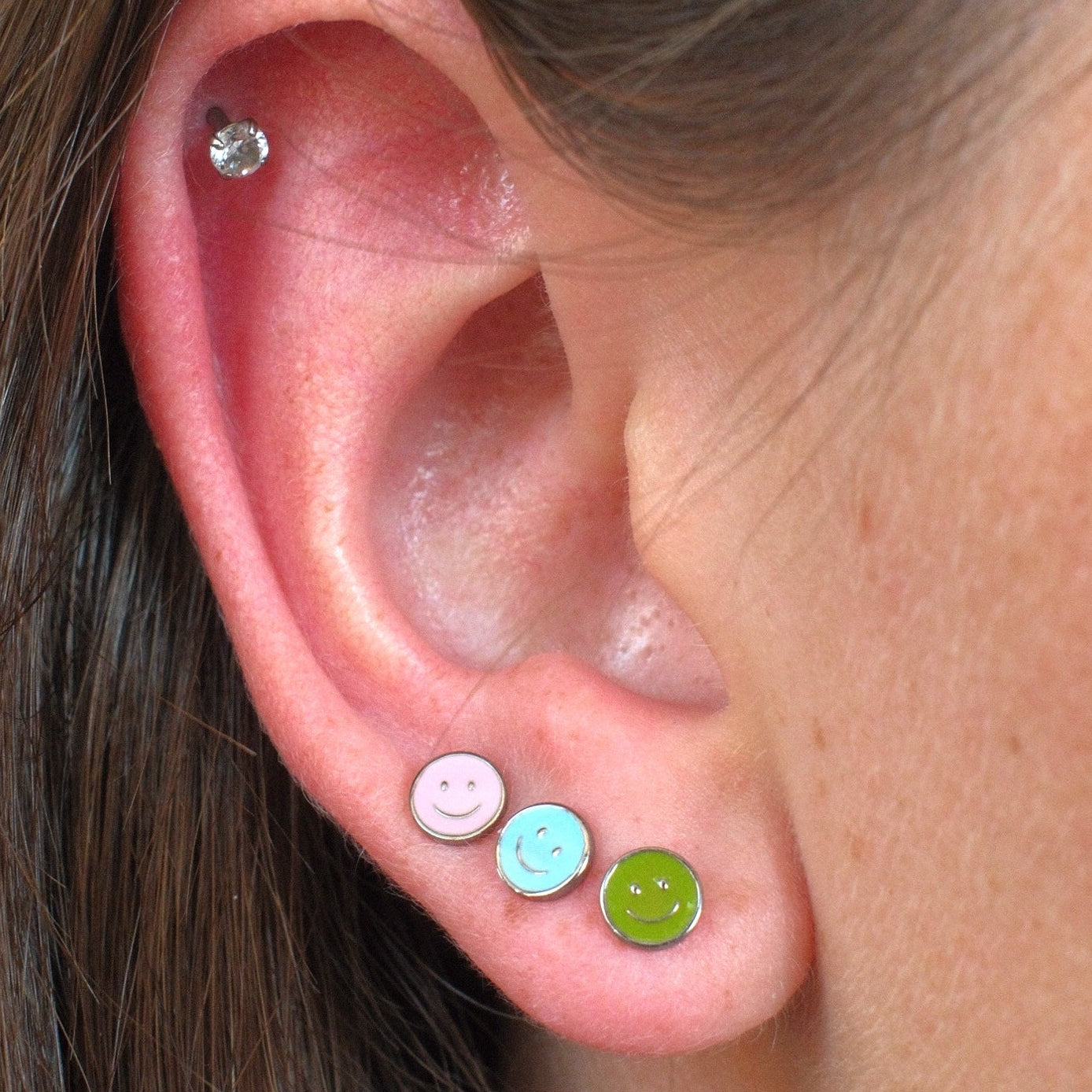 Close-up of a person's ear showcasing three colorful stud earrings: a pink smiley face stud, a light blue smiley face stud, and a lime green smiley face stud, with a small silver hoop earring in the upper ear cartilage.