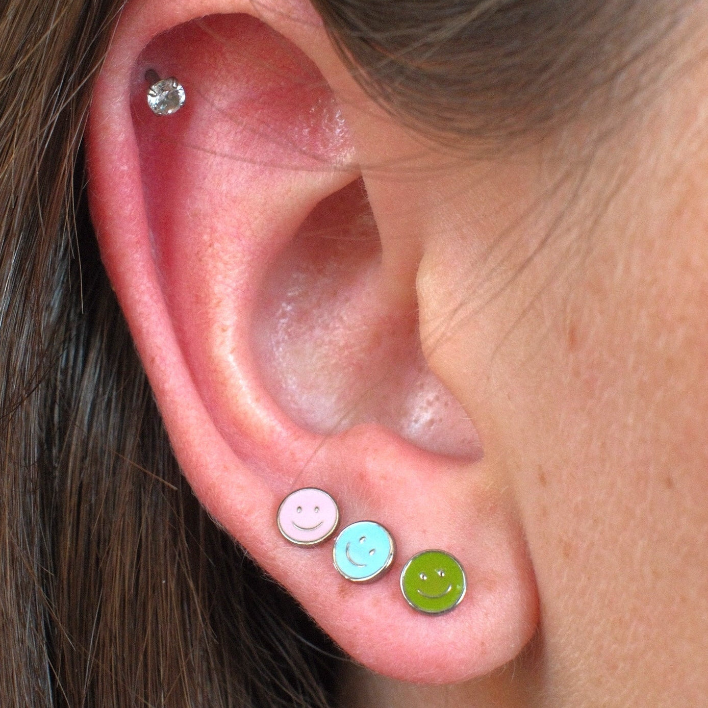 Close-up of a person's ear showcasing three colorful stud earrings: a pink smiley face stud, a light blue smiley face stud, and a lime green smiley face stud, with a small silver hoop earring in the upper ear cartilage.