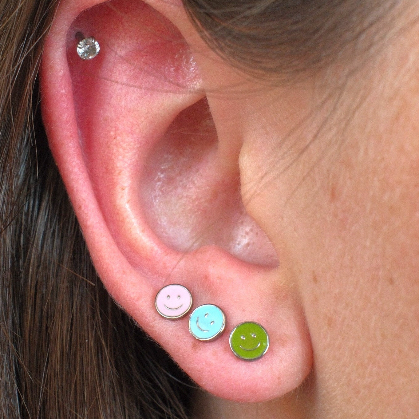 Close-up of a person's ear showcasing three colorful stud earrings: a pink smiley face stud, a light blue smiley face stud, and a lime green smiley face stud, with a small clear cz stud earring in the upper ear cartilage.