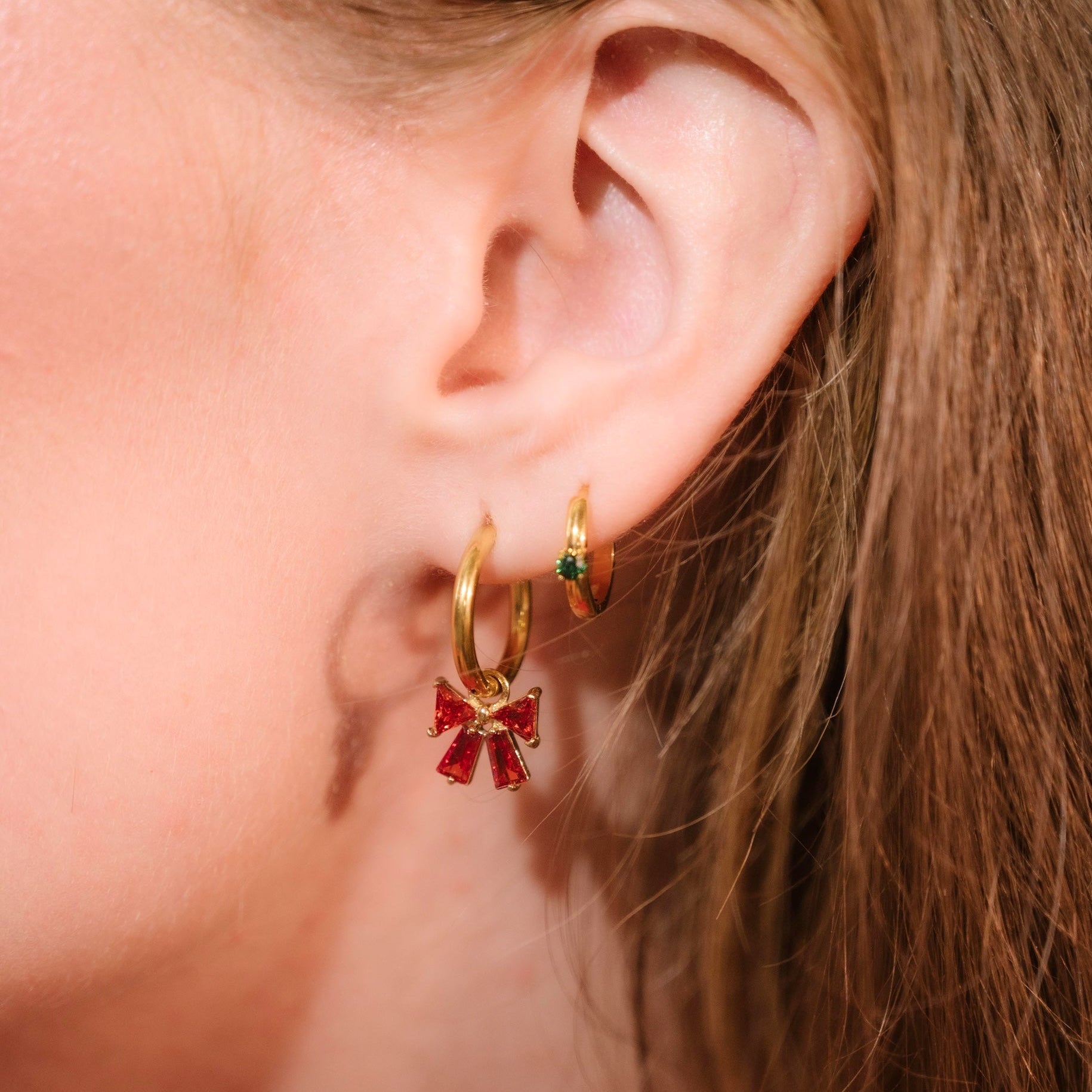 Close-up of a person wearing gold hoop earrings with red bow gem charm.