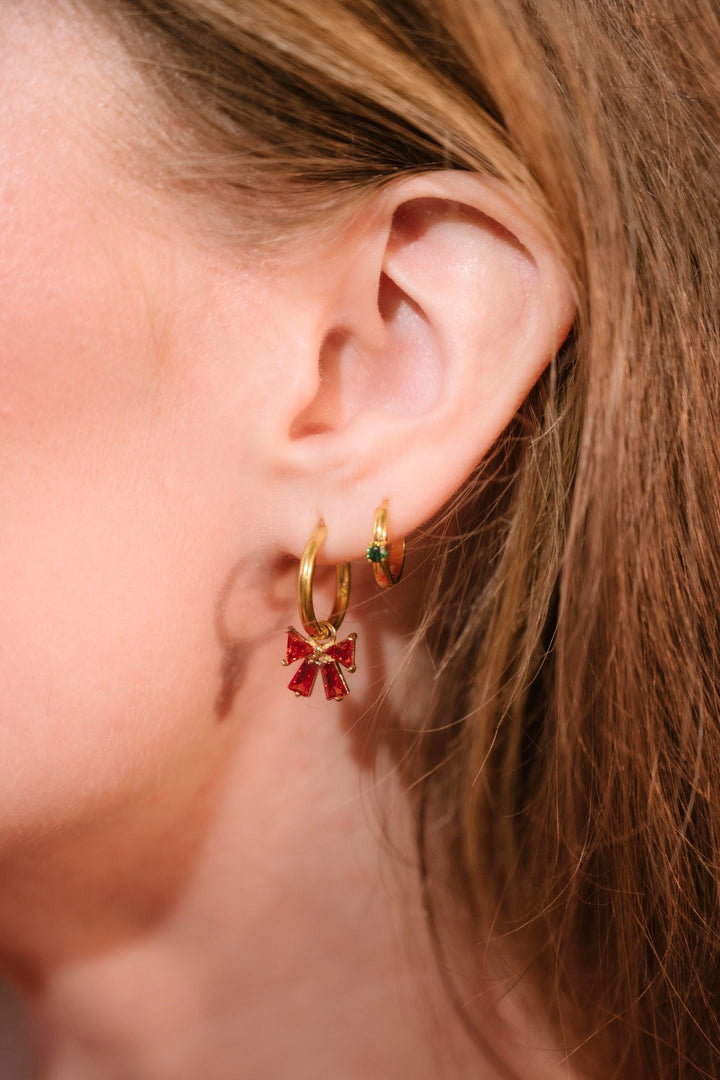 Close-up of a person wearing gold hoop earrings with red bow gem charm.