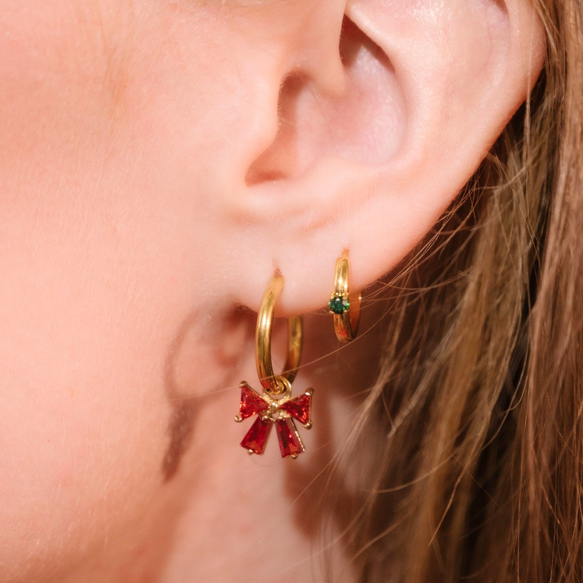 Close-up of an ear wearing gold hoop earrings with red and green gemstones.