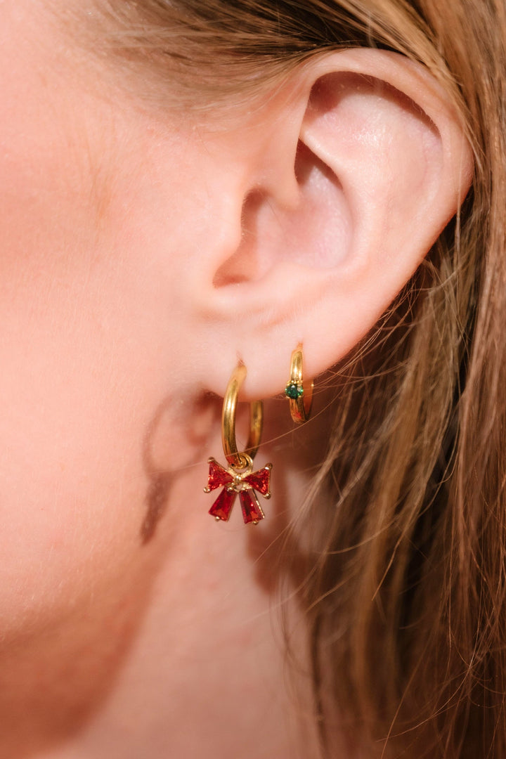 Close-up of an ear wearing gold hoop earrings with red and green gemstones.