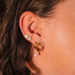 Close-up of a person's ear wearing gold hoop earrings with green gemstones.