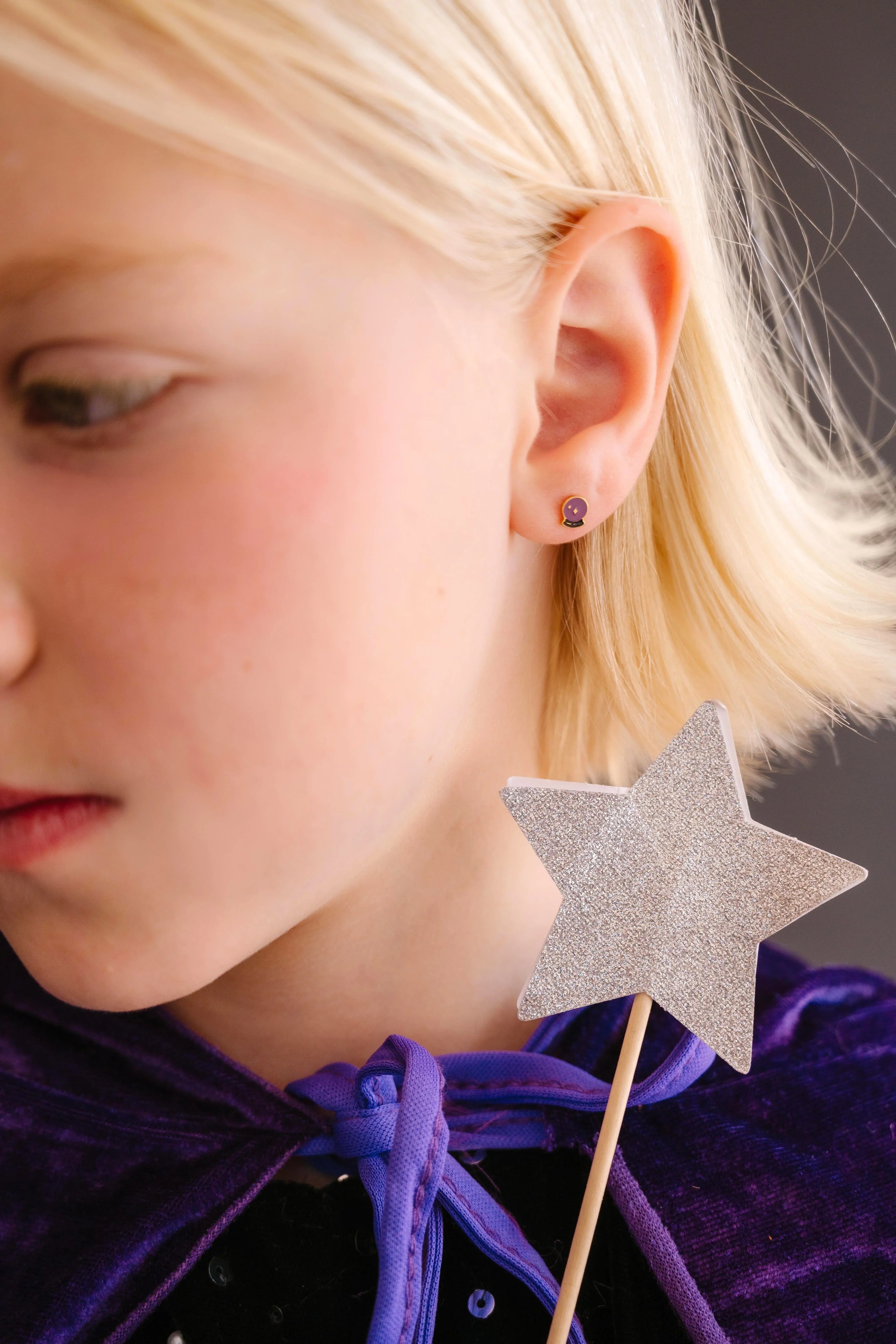 Close-up of child wearing a purple and black enamel flat back stud earring that is shaped like a crystal ball with star details within.