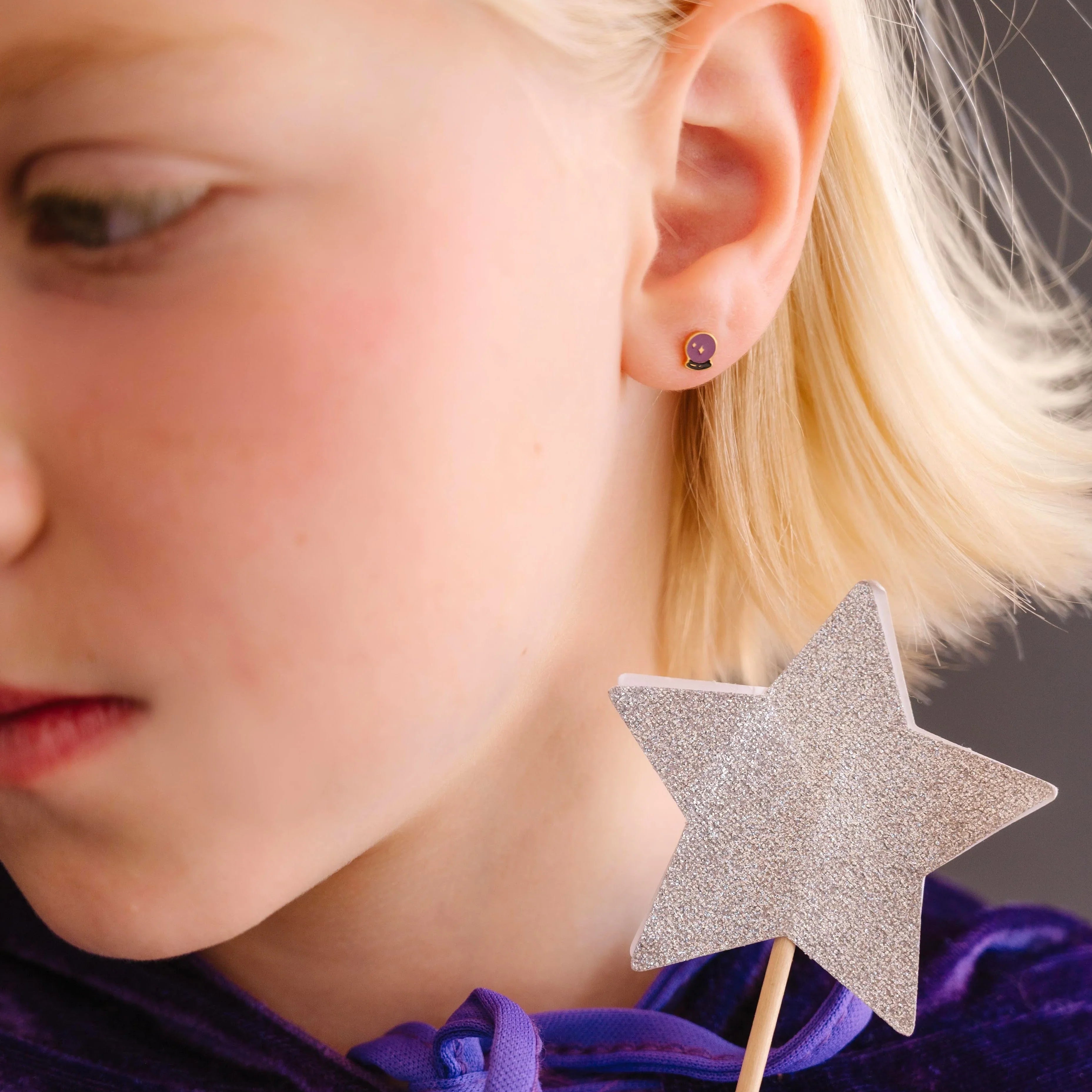 Close-up of child wearing a purple and black enamel flat back stud earring that is shaped like a crystal ball with star details within.