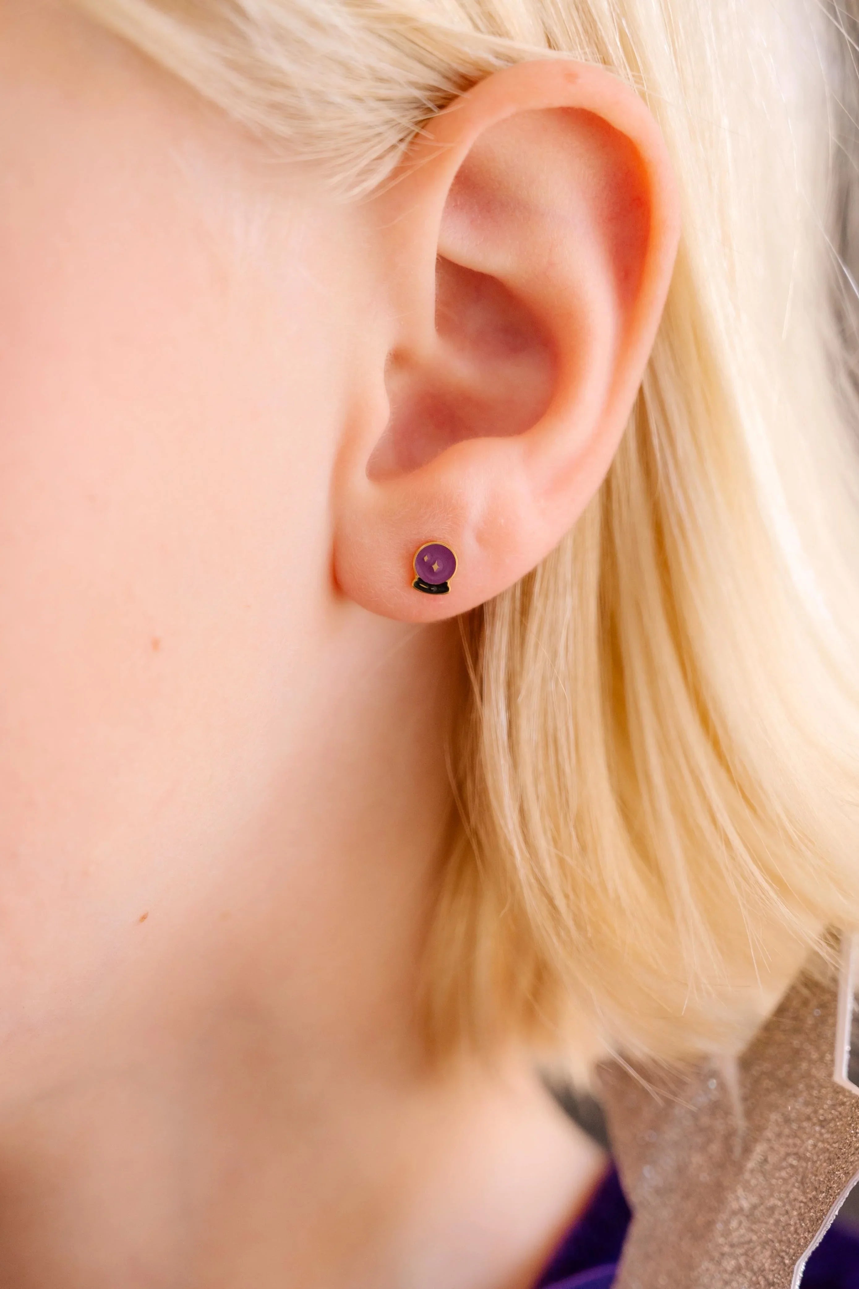 Close-up of an ear wearing a purple and black enamel flat back stud earring that is shaped like a crystal ball with star details within.