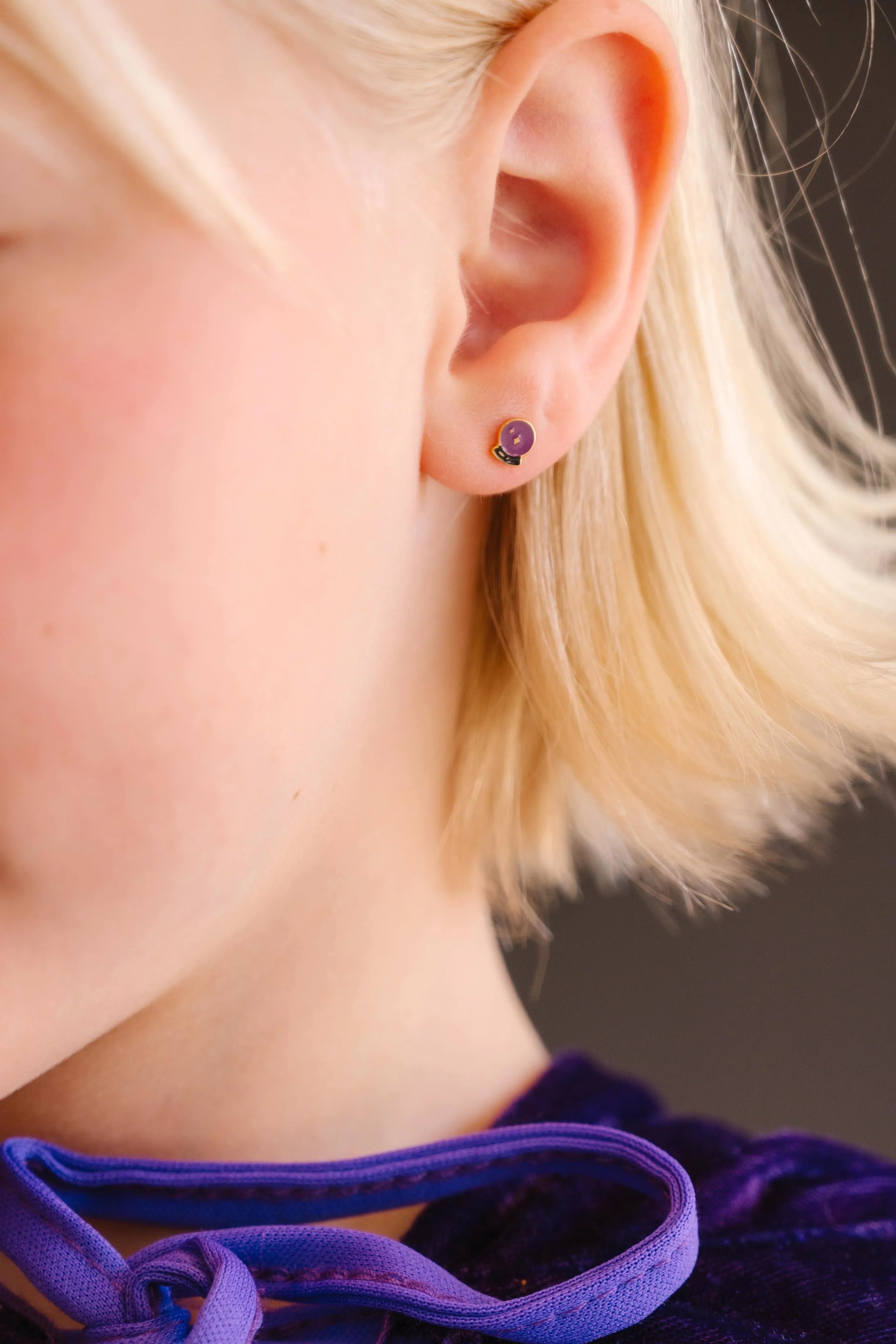 Close-up of an ear wearing a purple and black enamel flat back stud earring that is shaped like a crystal ball with star details within.