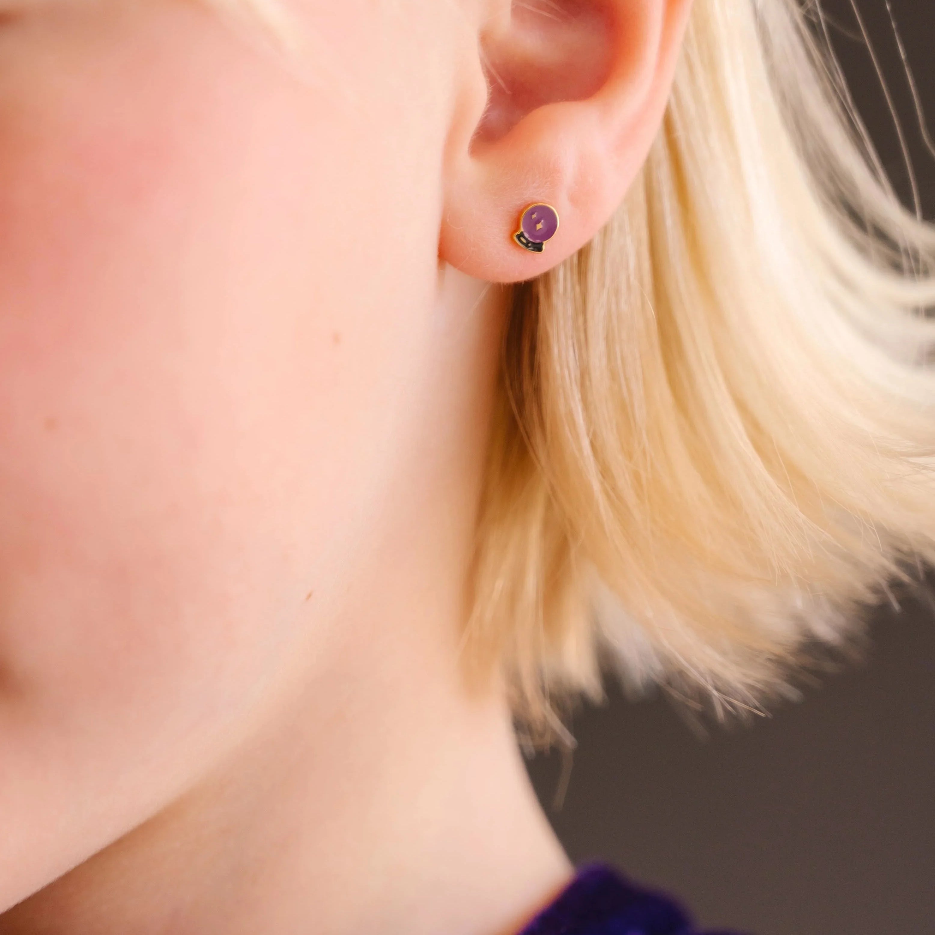 Close-up of an ear wearing a purple and black enamel flat back stud earring that is shaped like a crystal ball with star details within.