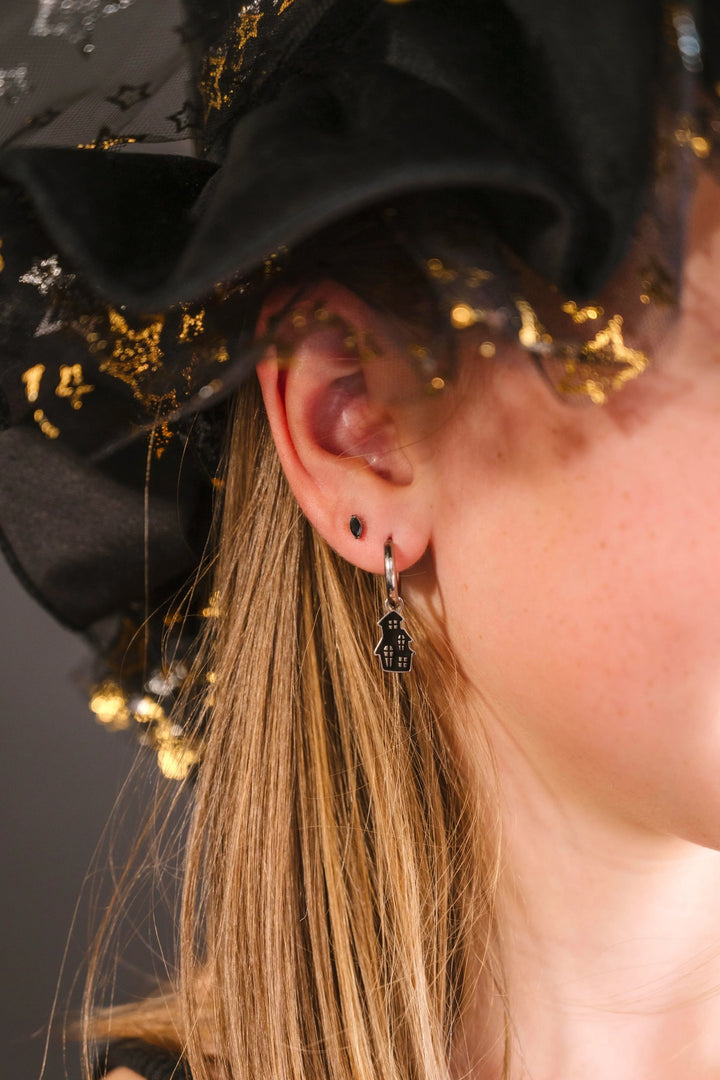 Close-up of an person wearing a black hat with gold star accents and wearing a silver hoop with a black enamel haunted house charm on it.