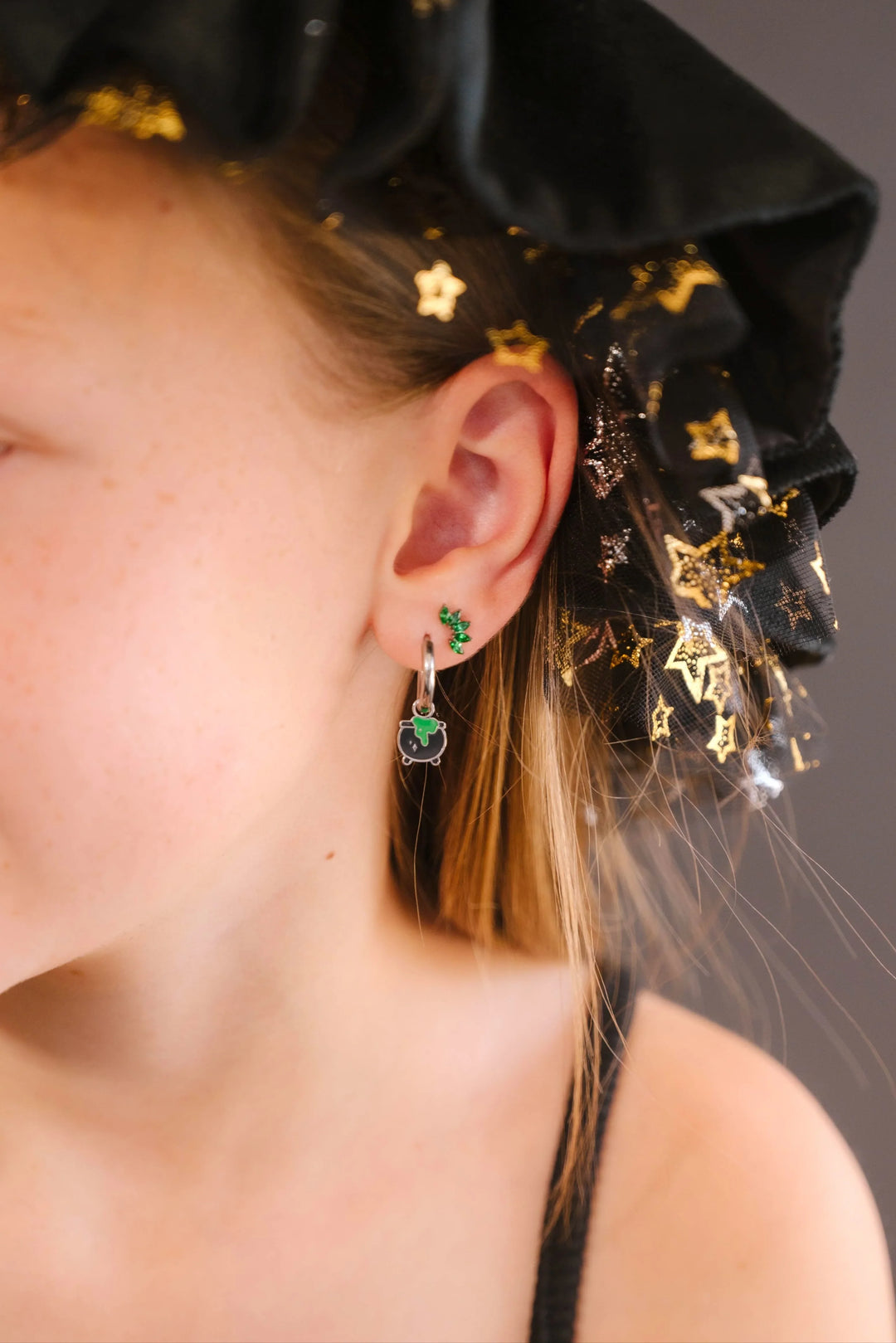 Close-up of a person in a black and gold starred witch hat, wearing a silver hoop with a black and green enamel cauldron charm.