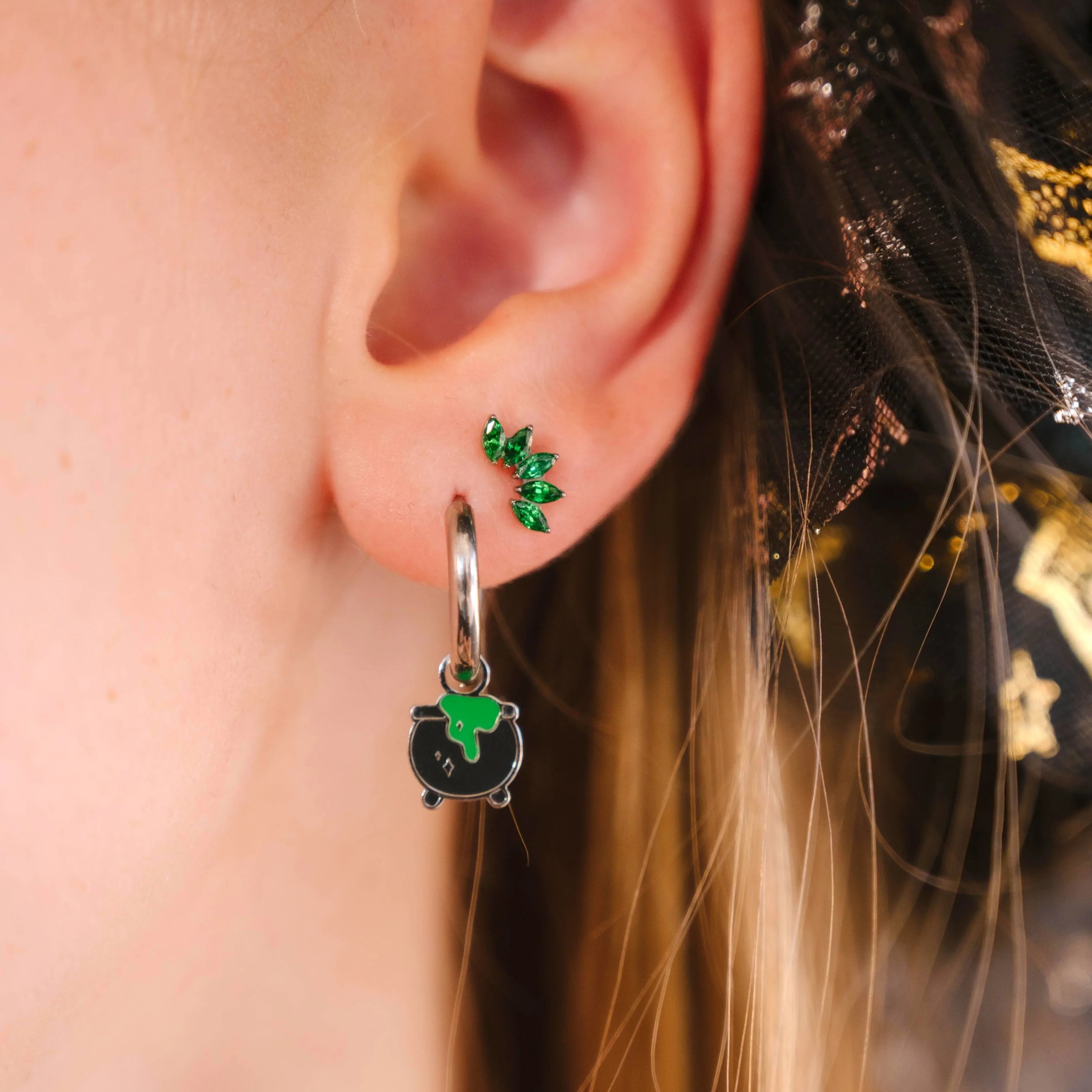 Close-up of an ear wearing a black and green enamel cauldron hoop charm with a blurred background
