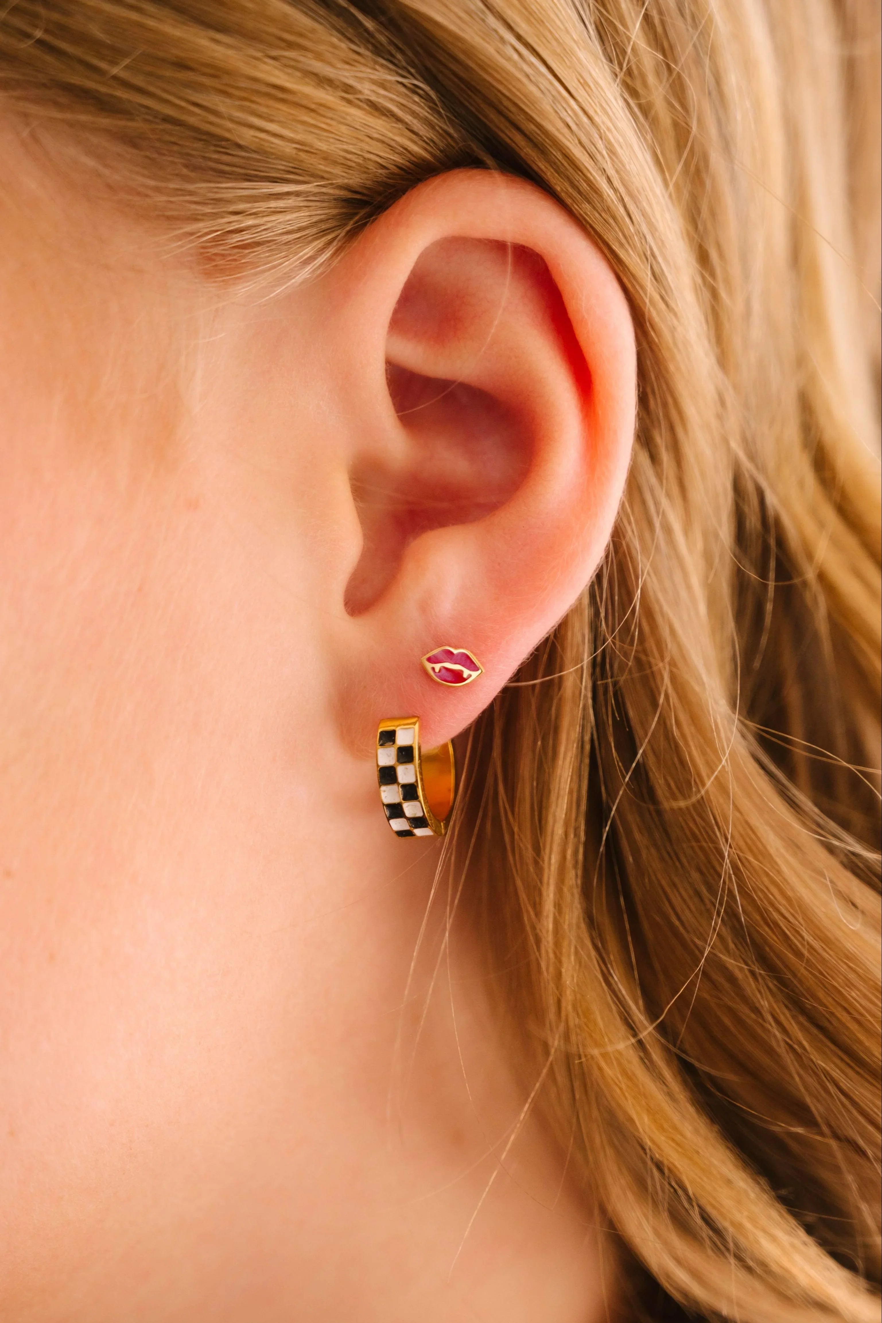 Close-up of an ear wearing a checkered hoop earring in a first lobe piercing and a red vampire lips stud in a second lobe piercing, with a blurred background