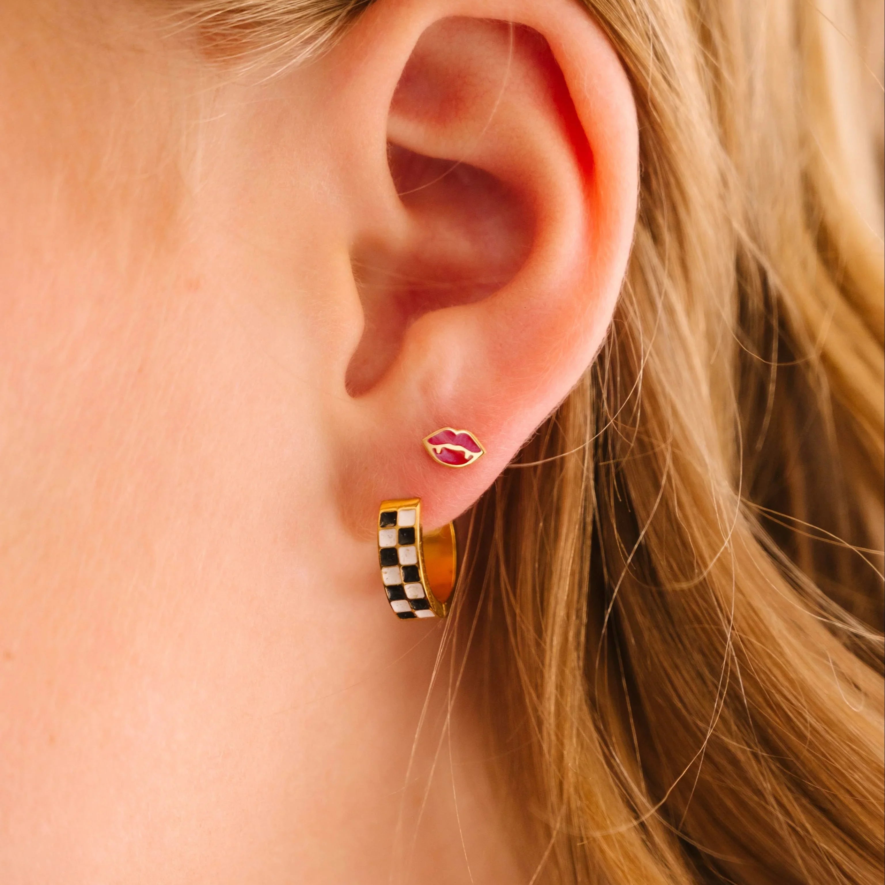 Close-up of an ear wearing a checkered hoop earring in a first lobe piercing and a red vampire lips stud in a second lobe piercing, with a blurred background