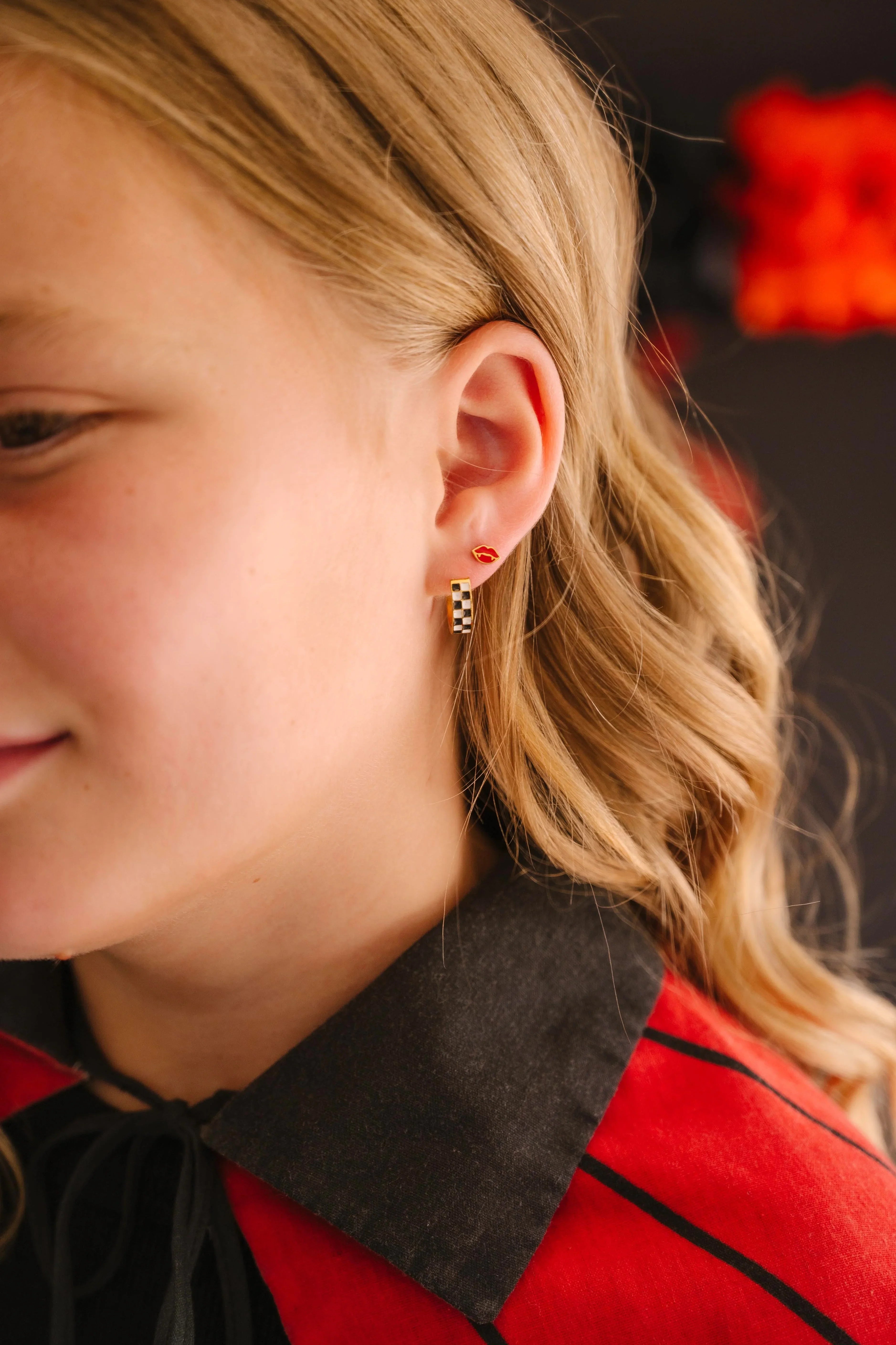 Close-up of a child wearing a red and black cloak, with a focus on the earrings in their ear, featuring a checkered hoop earring in a first lobe piercing and a red vampire lips stud in a second lobe piercing, with a blurred background