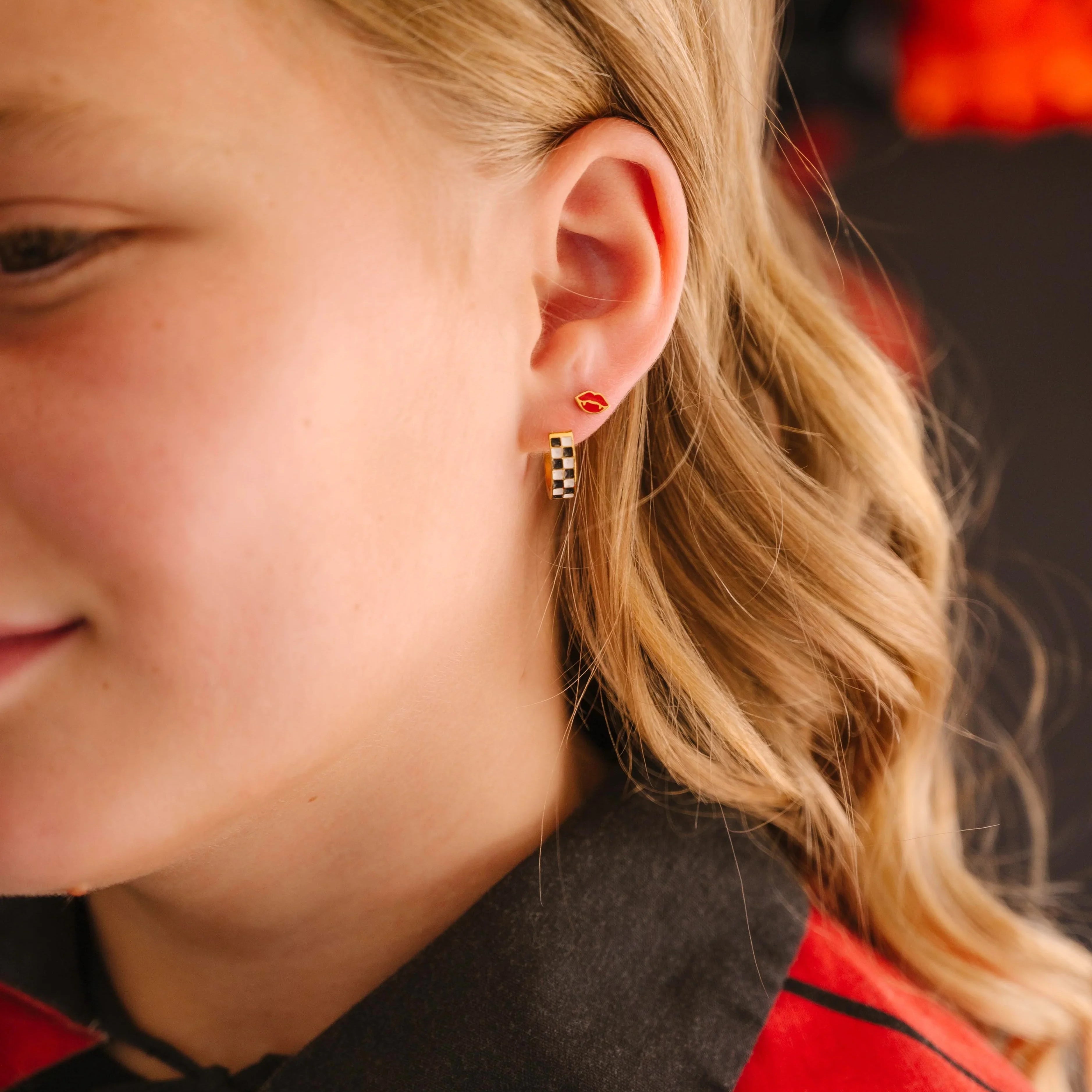Close-up of a child wearing a red and black cloak, with a focus on the earrings in their ear, featuring a checkered hoop earring in a first lobe piercing and a red vampire lips stud in a second lobe piercing, with a blurred background