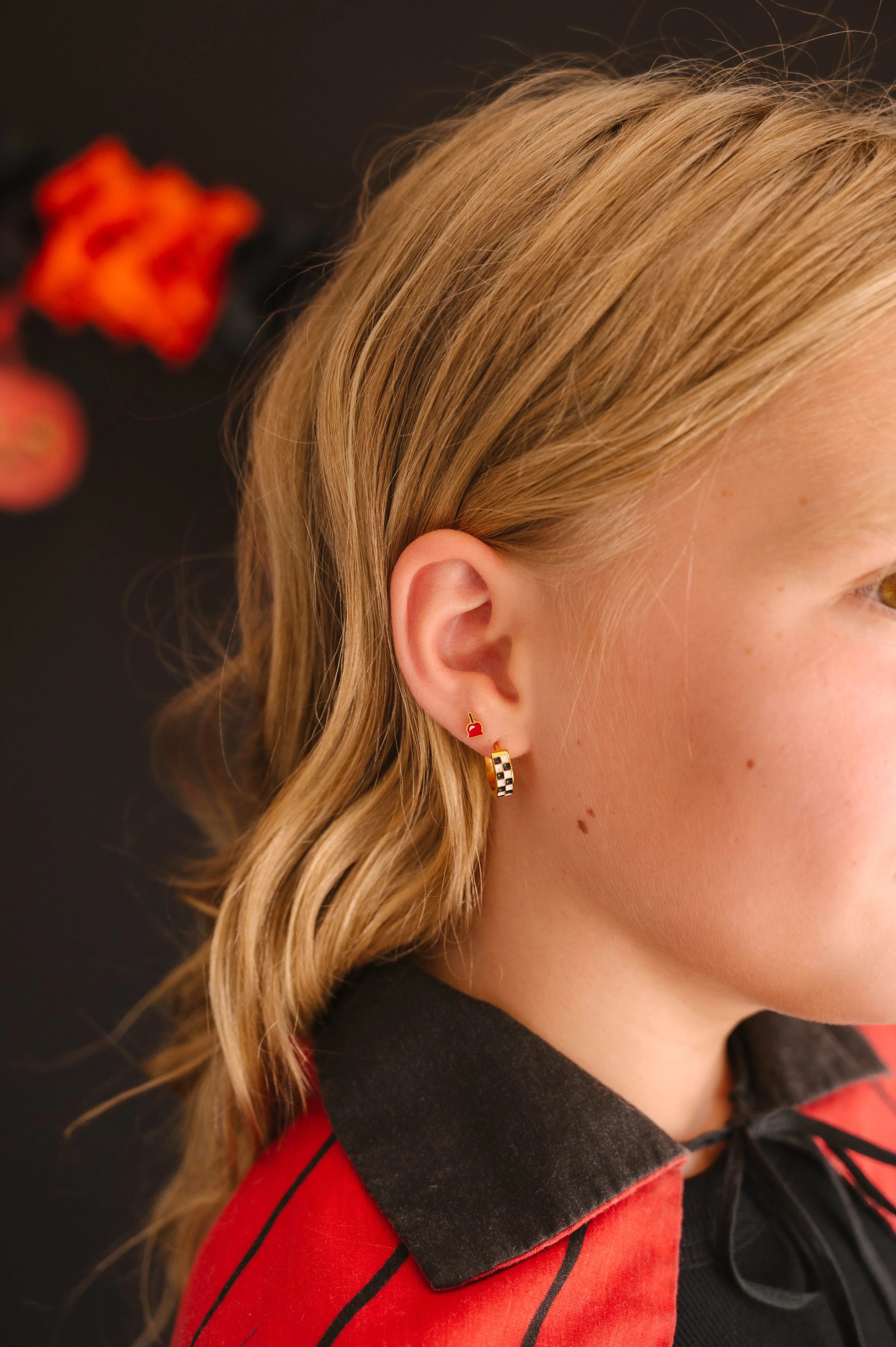 Close-up of a person wearing a red cloak and focusing on their ear, featuring a checkered hoop earring in a first lobe piercing and a bright red enamel stud shaped like a candied apple in a second lobe piercing.