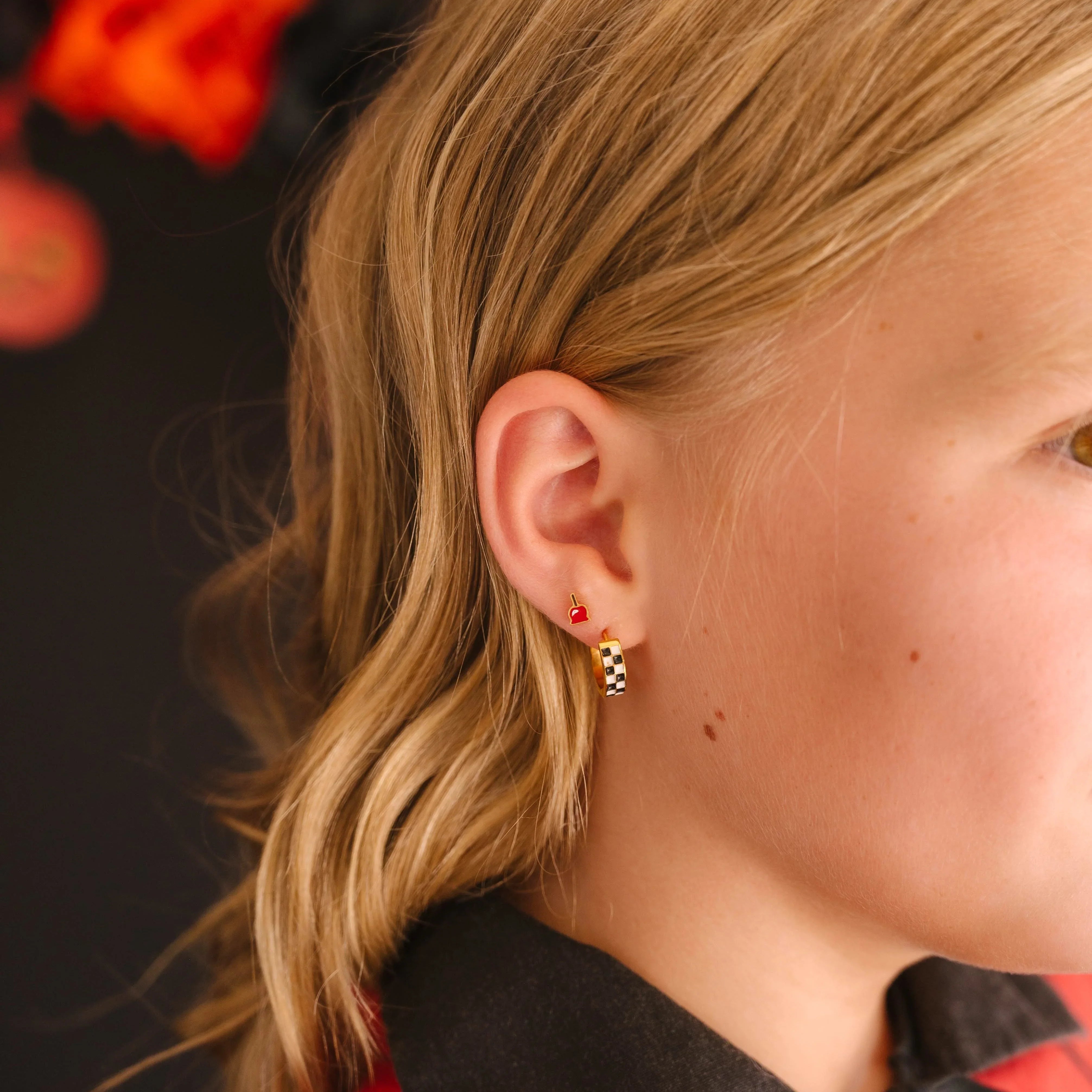 Close-up of a person wearing a red cloak and focusing on their ear, featuring a checkered hoop earring in a first lobe piercing and a bright red enamel stud shaped like a candied apple in a second lobe piercing.